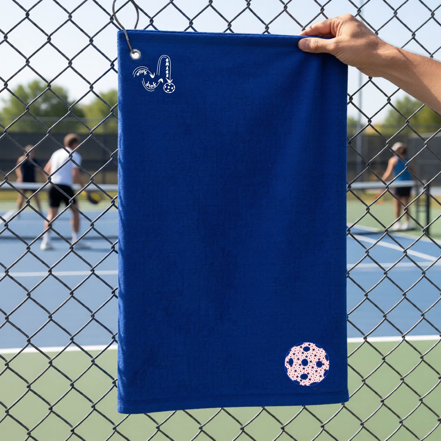 A royal blue terry cloth court towel is held up in front of a chain-link fence at a pickleall court. The towel features a silver metal grommet and hook at the top, with the text"DINK DINK SMASH" and a colorful pickleball graphic near the bottom right.