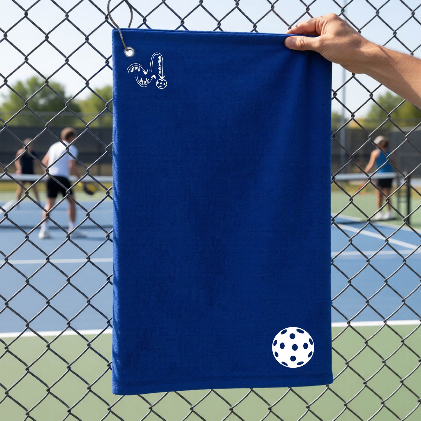 A royal blue terry cloth court towel is held up in front of a chain-link fence at a pickleall court. The towel features a silver metal grommet and hook at the top, with the text"DINK DINK SMASH" and a colorful pickleball graphic near the bottom right.