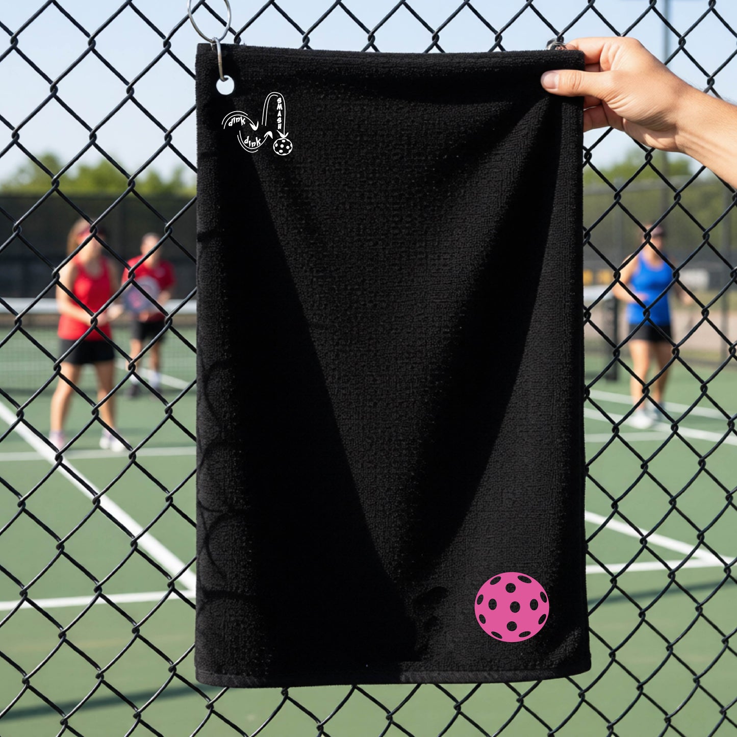 A black terry cloth towel is held up in front of a chain-link fence at a pickleall court. The towel features a silver metal grommet and hook at the top, with the text"DINK DINK SMASH" and a colorful pickleball graphic near the bottom right.