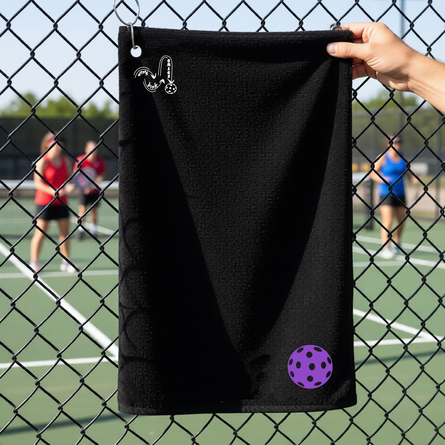 A black terry cloth towel is held up in front of a chain-link fence at a pickleall court. The towel features a silver metal grommet and hook at the top, with the text"DINK DINK SMASH" and a colorful pickleball graphic near the bottom right.
