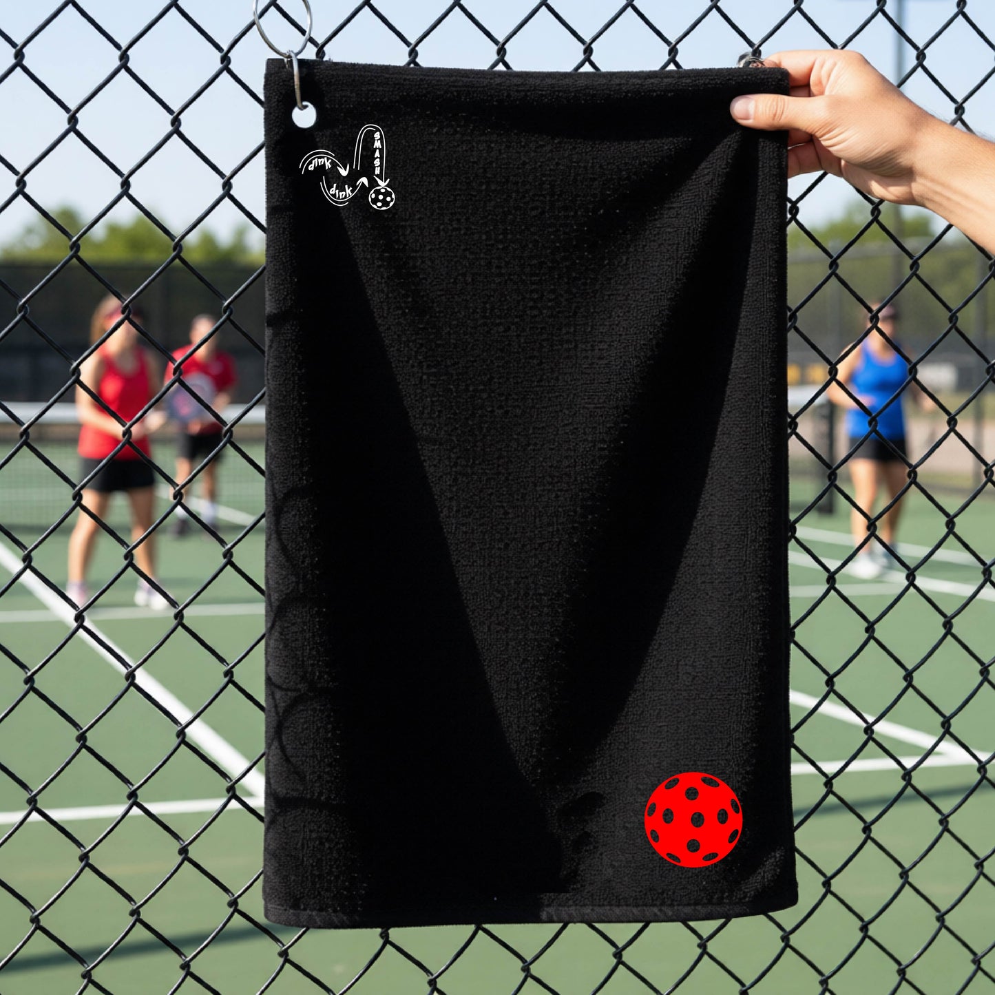 A black terry cloth court towel is held up in front of a chain-link fence at a pickleall court. The towel features a silver metal grommet and hook at the top, with the text"DINK DINK SMASH" and a colorful pickleball graphic near the bottom right.