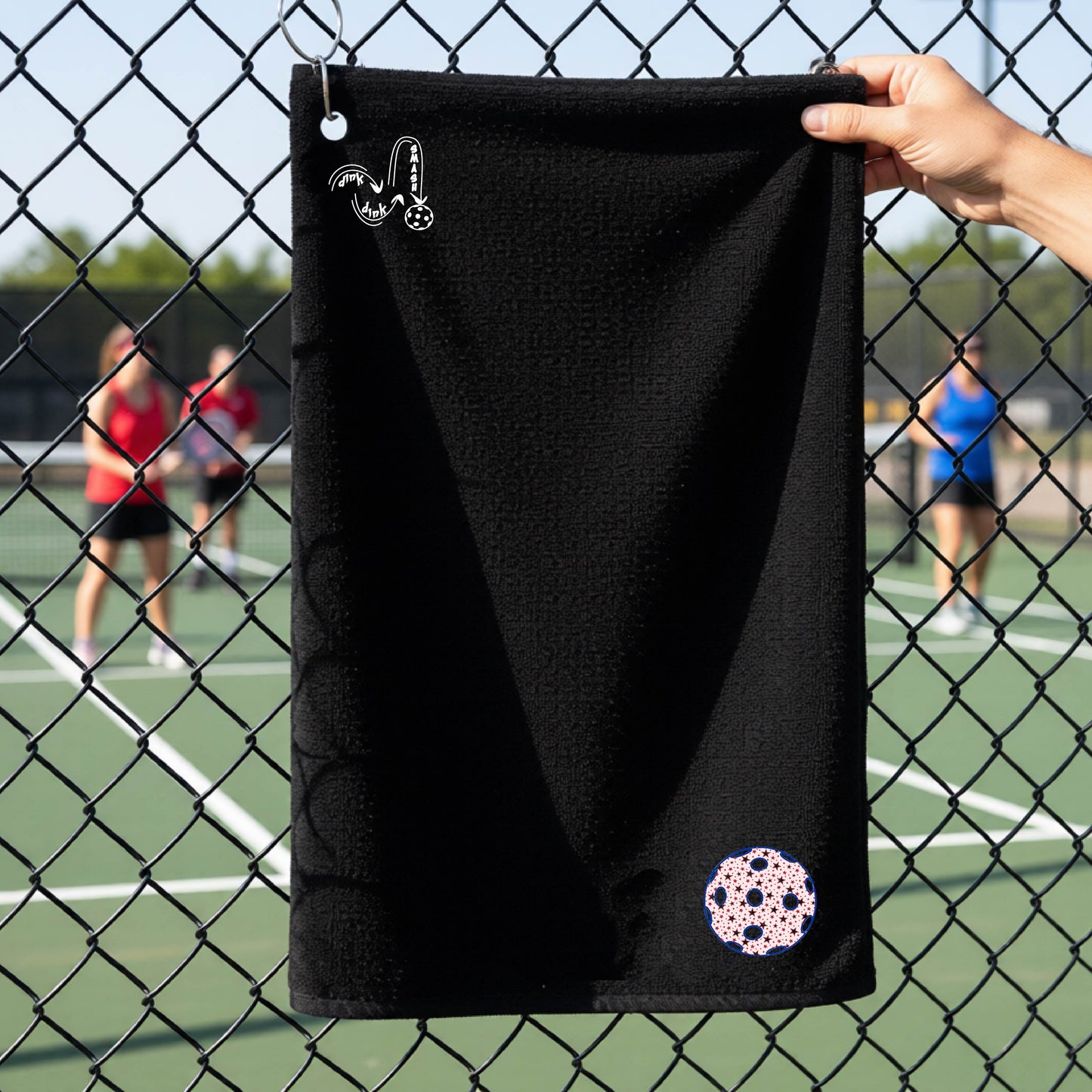 A black terry cloth court towel is held up in front of a chain-link fence at a pickleall court. The towel features a silver metal grommet and hook at the top, with the text"DINK DINK SMASH" and a colorful pickleball graphic near the bottom right.