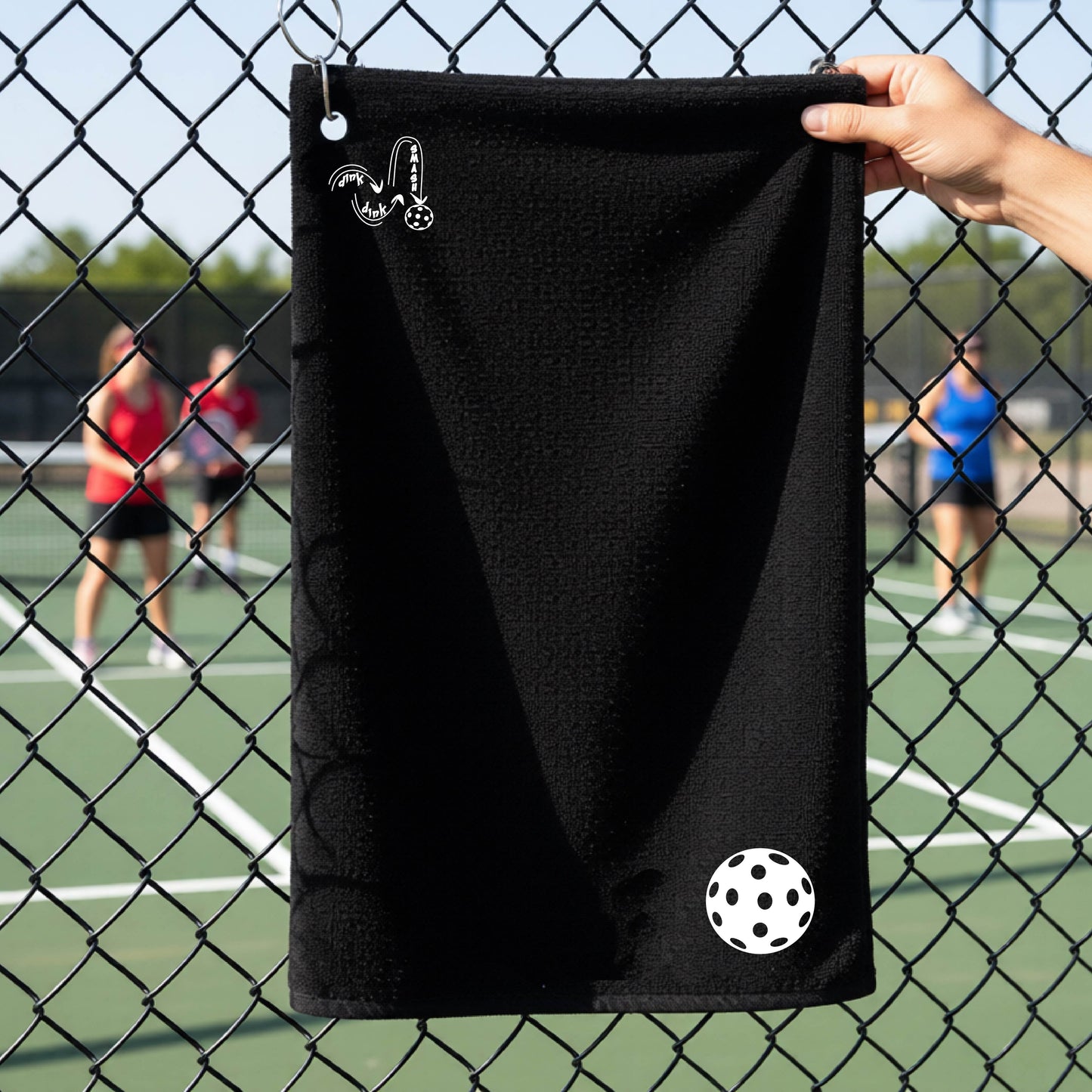 A black terry cloth court towel is held up in front of a chain-link fence at a pickleall court. The towel features a silver metal grommet and hook at the top, with the text"DINK DINK SMASH" and a colorful pickleball graphic near the bottom right.
