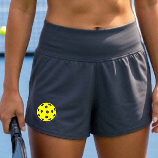 Gray athletic shorts with side pockets, shown on a model on a pickleball court. The shorts feature a colorful pickleball graphic on the left thigh.