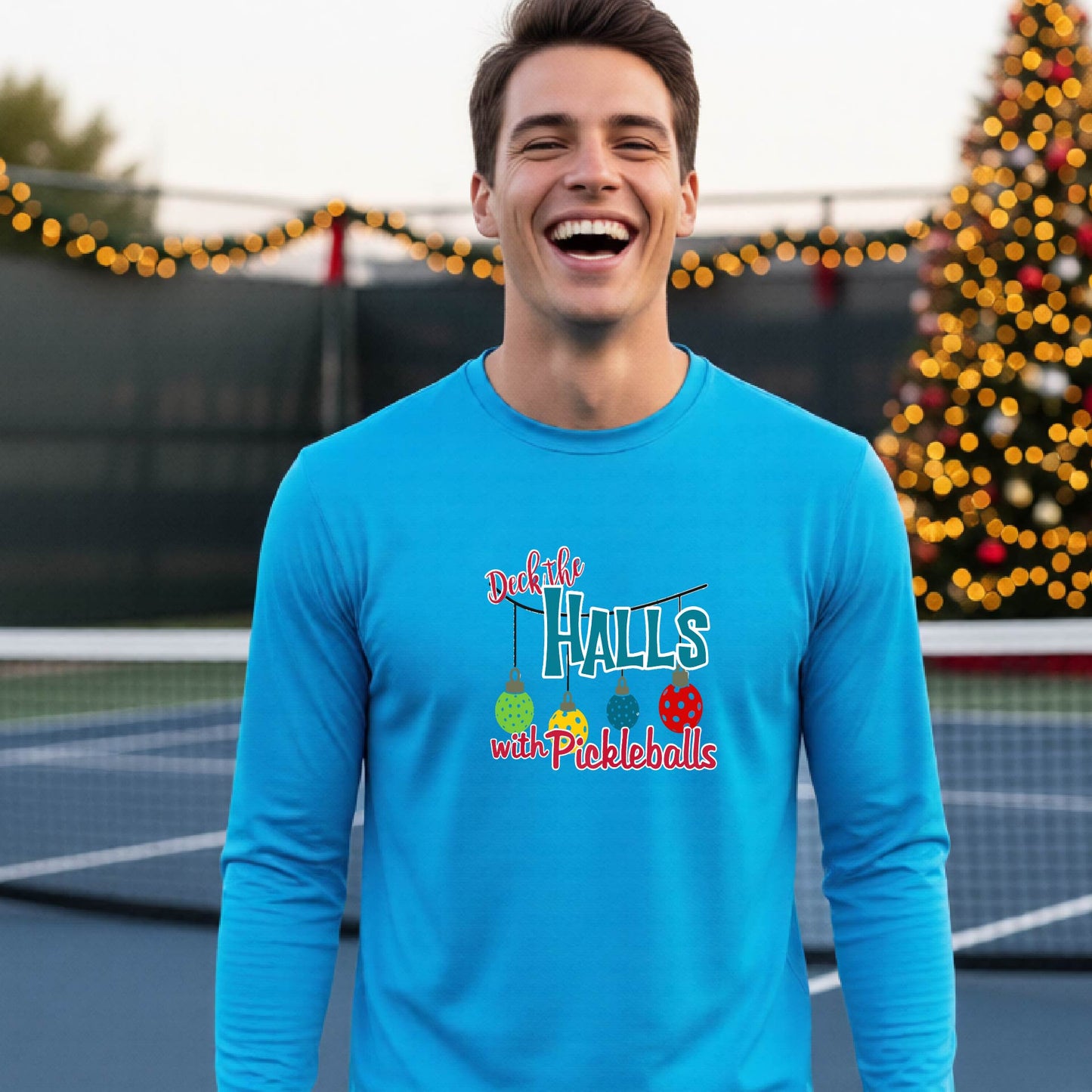 A smiling man wearing a light blue long-sleeve t-shirt with a colorful graphic that reads 'Deck the Halls with Pickleballs' and holiday-themed graphics, standing on an outdoor pickleball court with festive lights in the background.