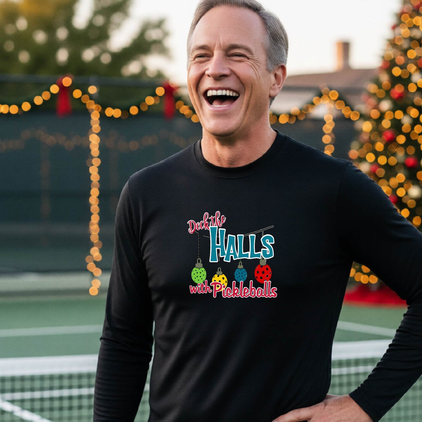 A smiling man wearing a black long-sleeve t-shirt with a colorful graphic that reads 'Deck the Halls with Pickleballs' and holiday-themed graphics, standing on an outdoor pickleball court with festive lights in the background.