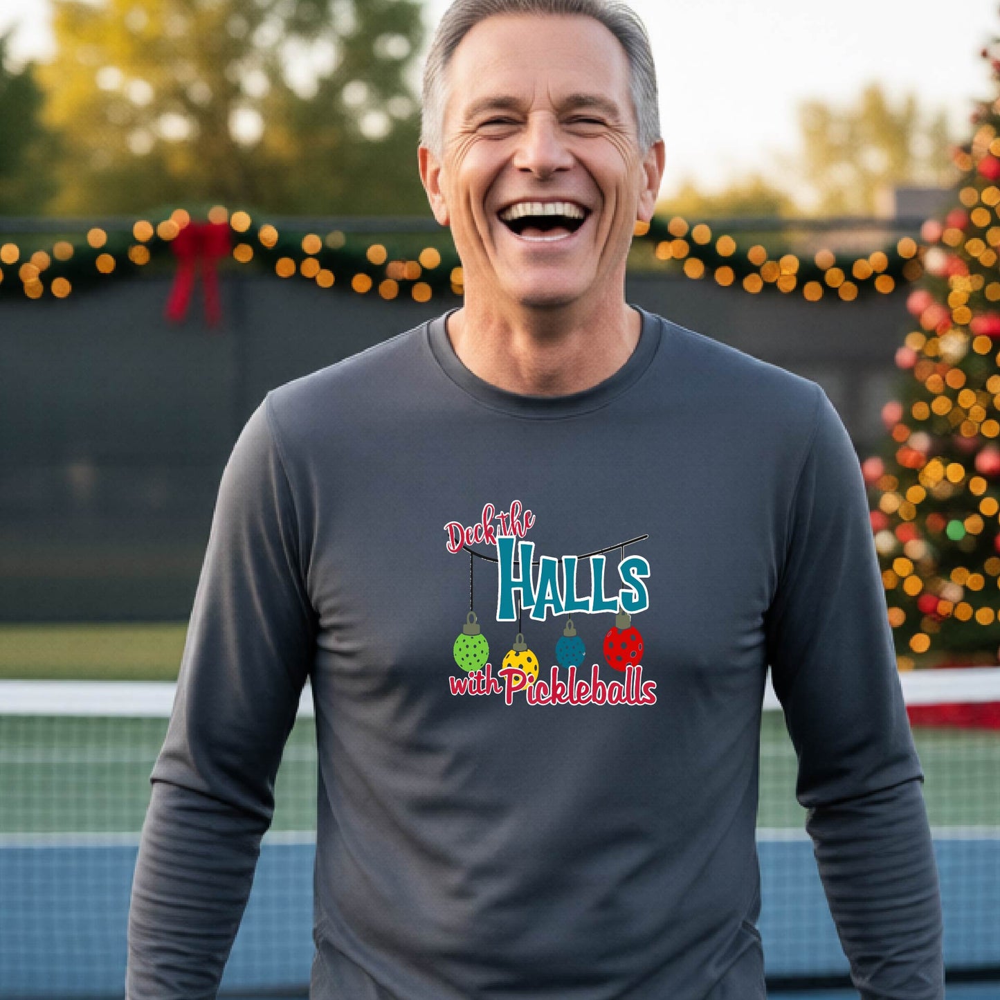 A smiling man wearing a dark gray long-sleeve t-shirt with a colorful graphic that reads 'Deck the Halls with Pickleballs' and holiday-themed graphics, standing on an outdoor pickleball court with festive lights in the background.