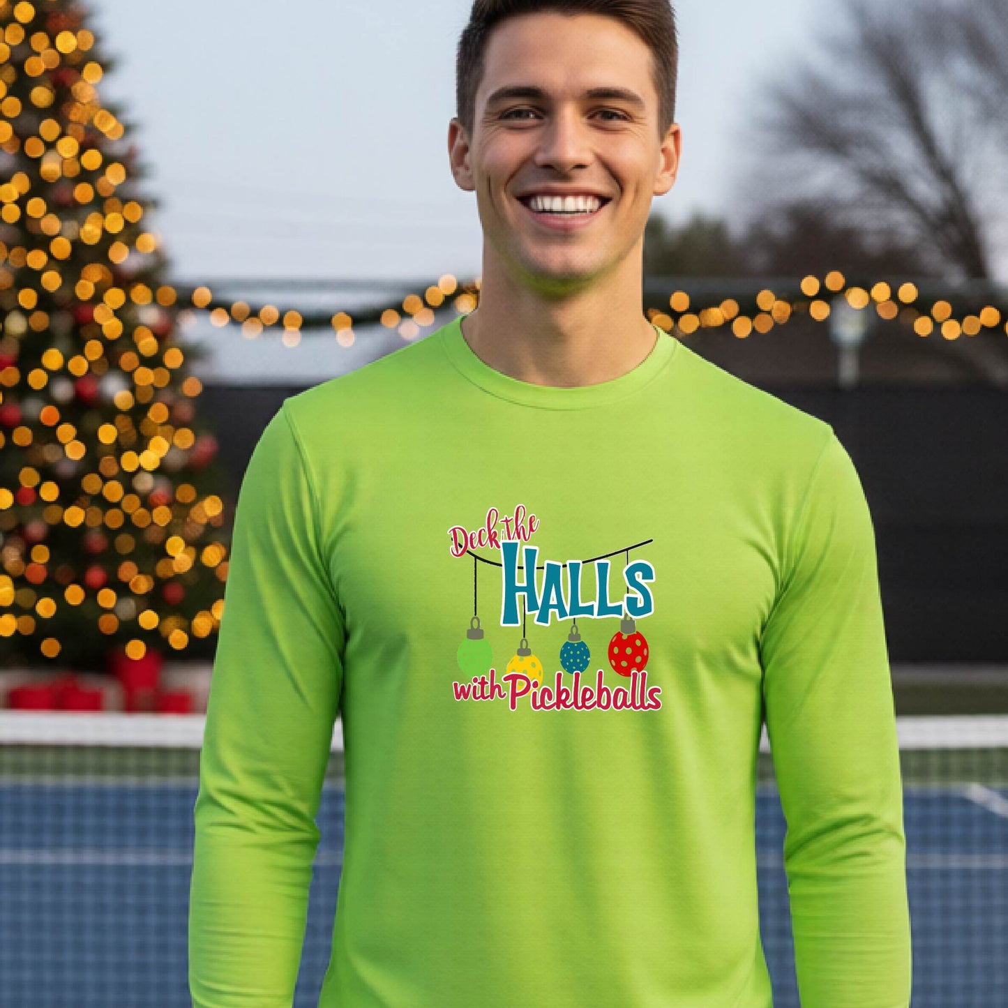 A smiling man wearing a lime green long-sleeve t-shirt with a colorful graphic that reads 'Deck the Halls with Pickleballs' and holiday-themed graphics, standing on an outdoor pickleball court with festive lights in the background.