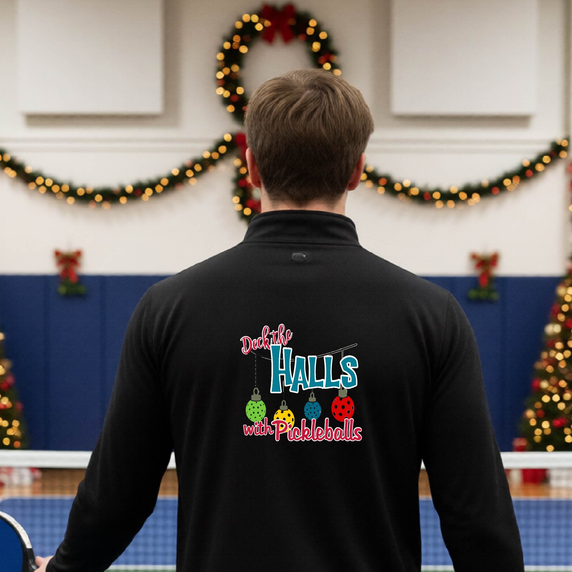 An man viewed from behind wearing a black quarter-zip pullover with a 'Deck the Halls with Pickleballs' graphic on the back, standing in a gym decorated for Christmas.