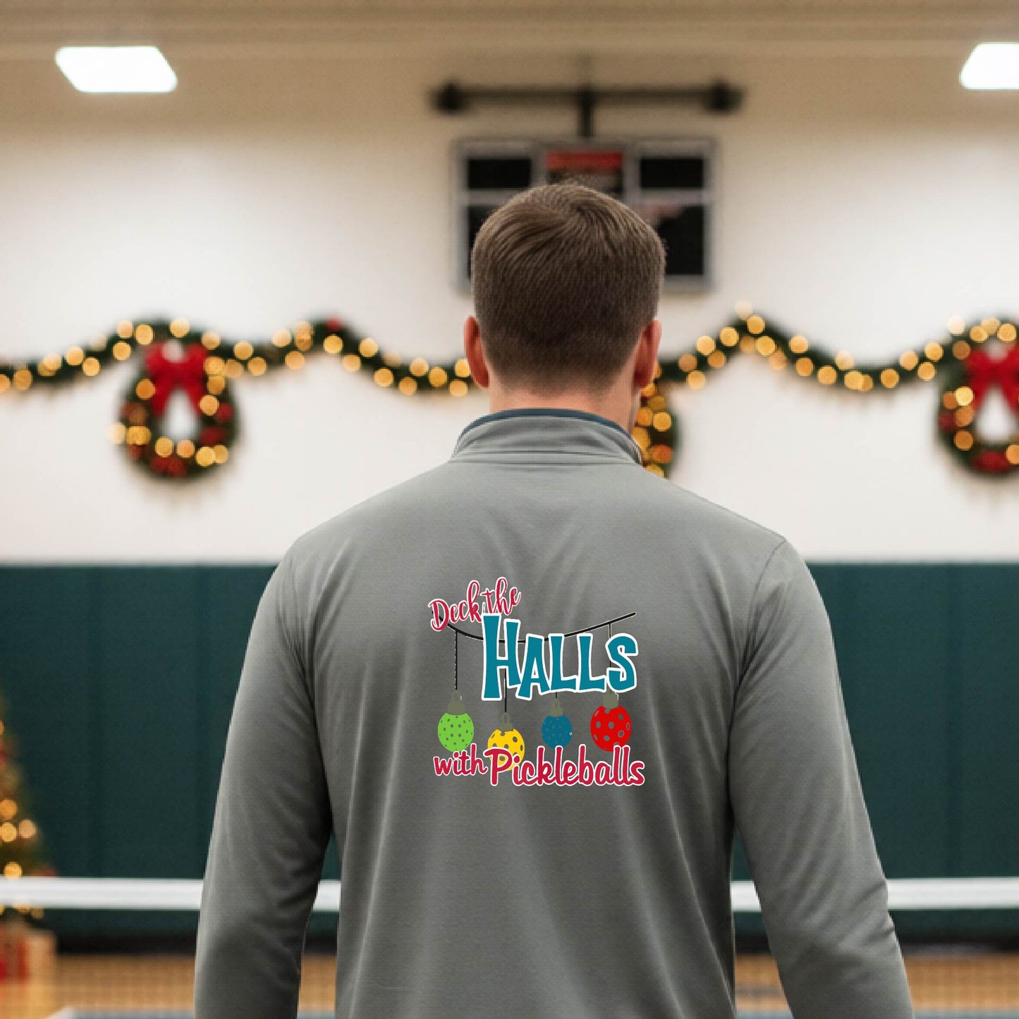An man viewed from behind wearing a gray quarter-zip pullover with a 'Deck the Halls with Pickleballs' graphic on the back, standing in a gym decorated for Christmas.