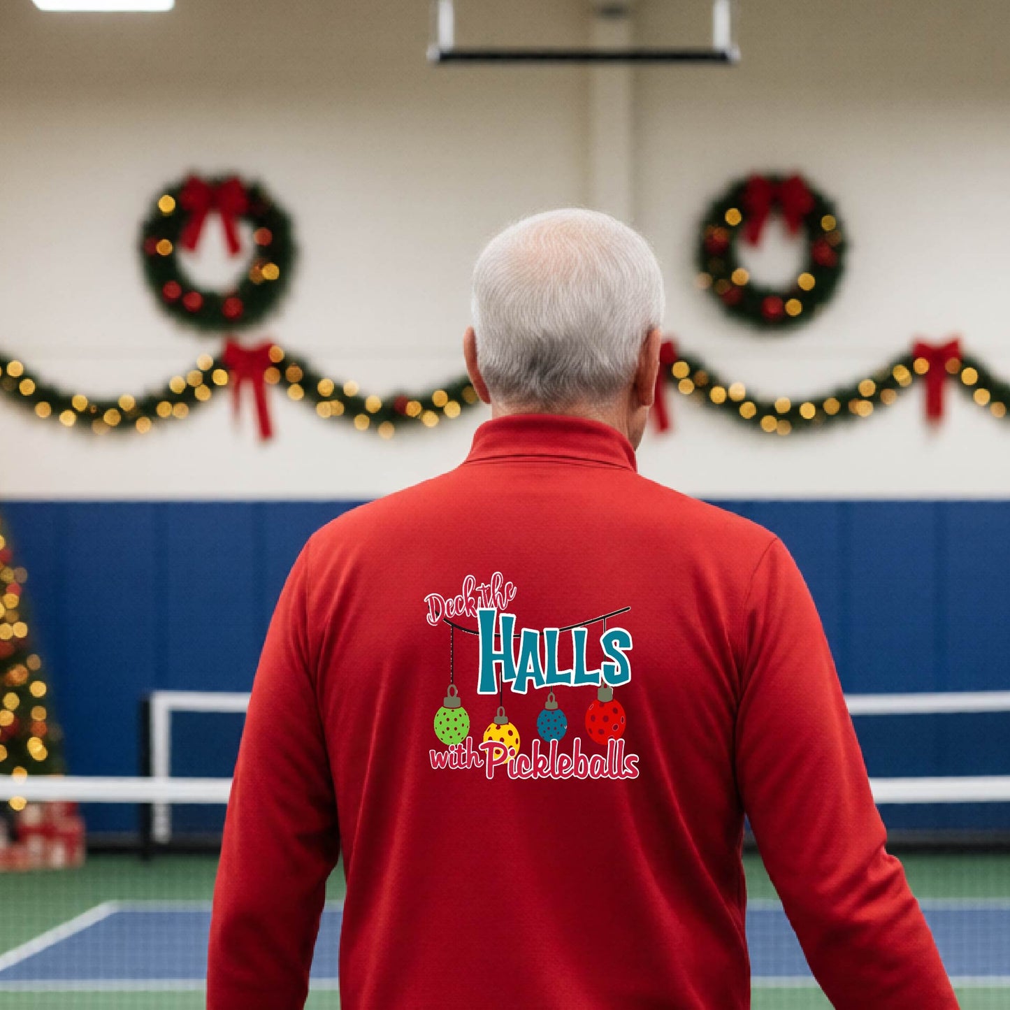 An man viewed from behind wearing a red quarter-zip pullover with a 'Deck the Halls with Pickleballs' graphic on the back, standing in a gym decorated for Christmas.