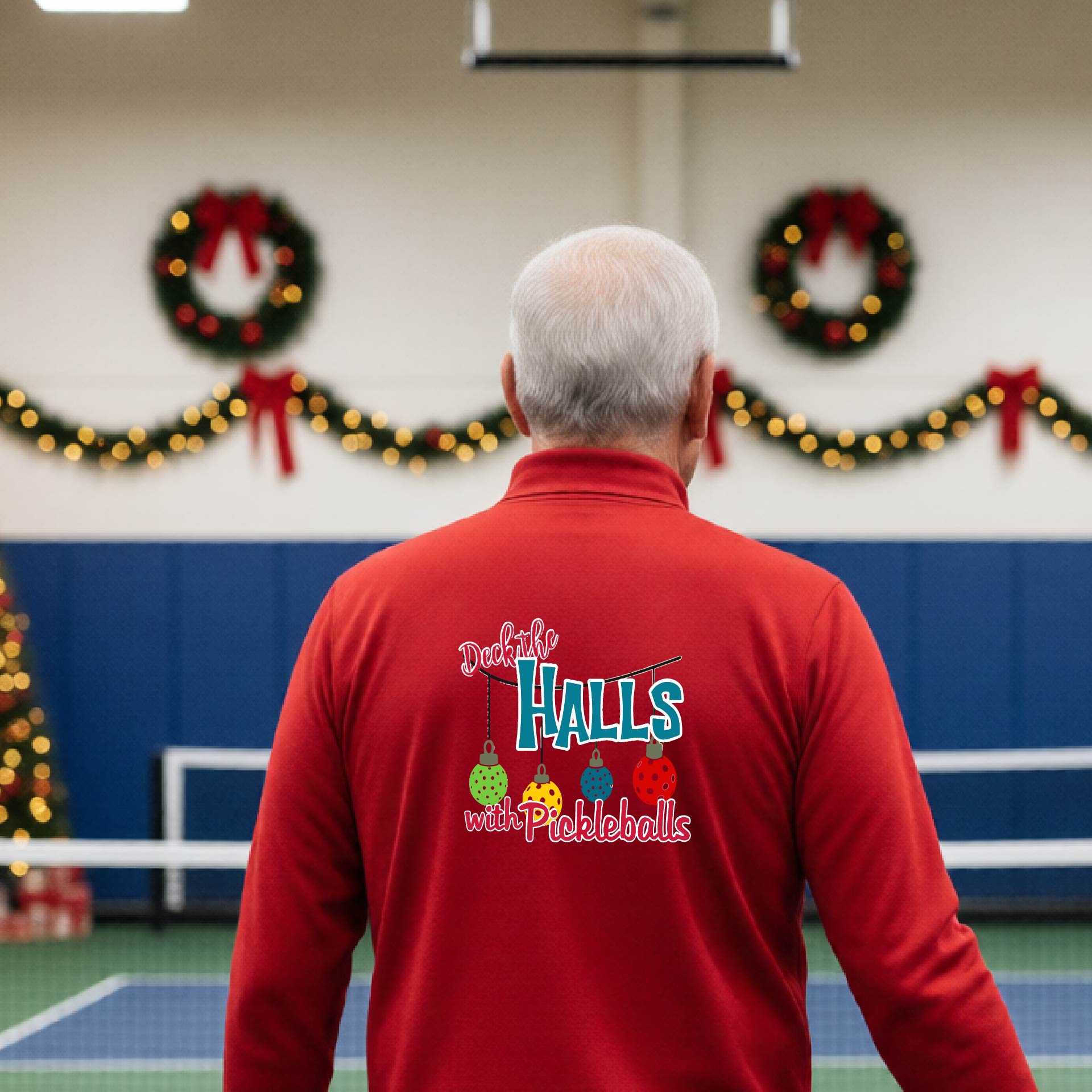 An man viewed from behind wearing a red quarter-zip pullover with a 'Deck the Halls with Pickleballs' graphic on the back, standing in a gym decorated for Christmas.