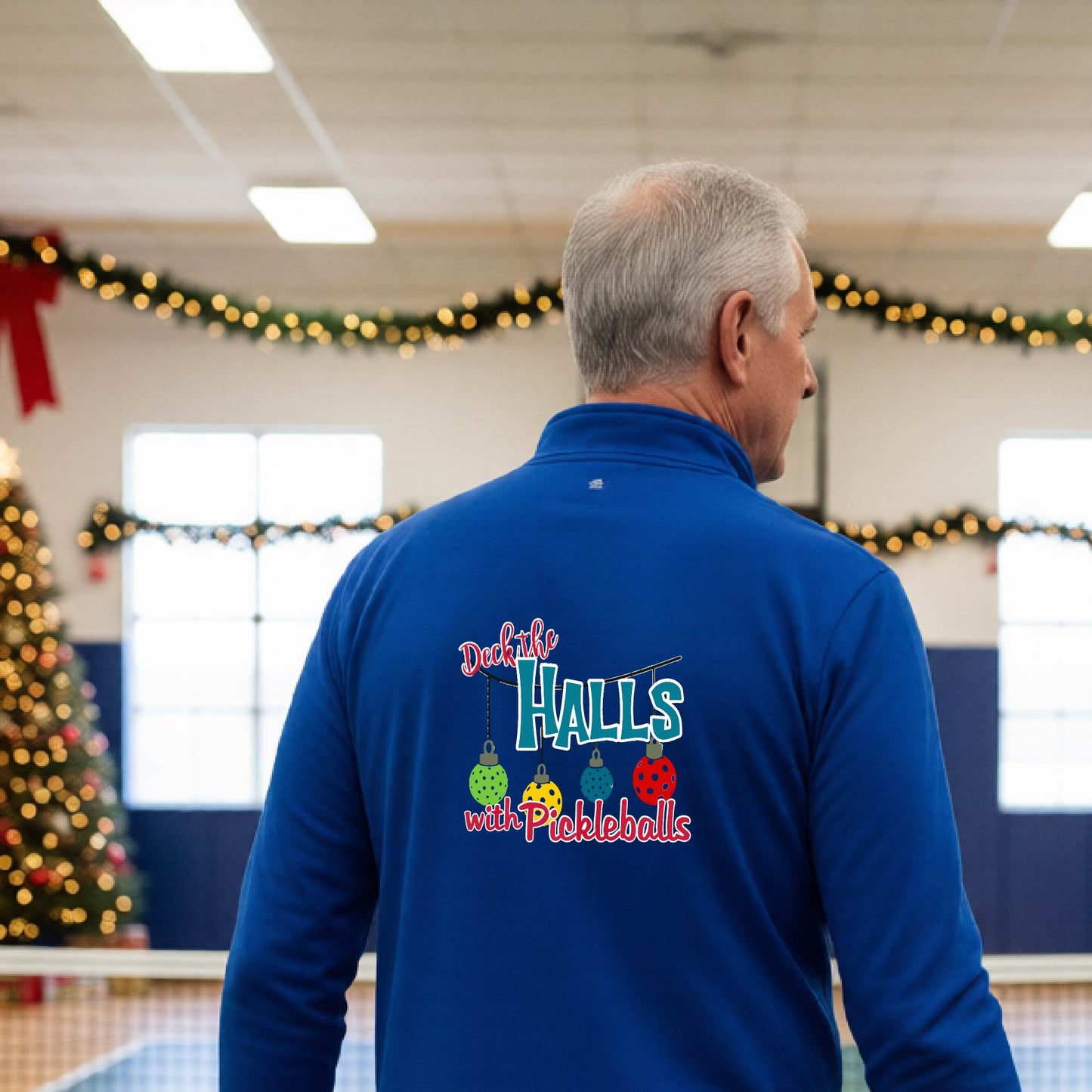 An man viewed from behind wearing a royal blue quarter-zip pullover with a 'Deck the Halls with Pickleballs' graphic on the back, standing in a gym decorated for Christmas.