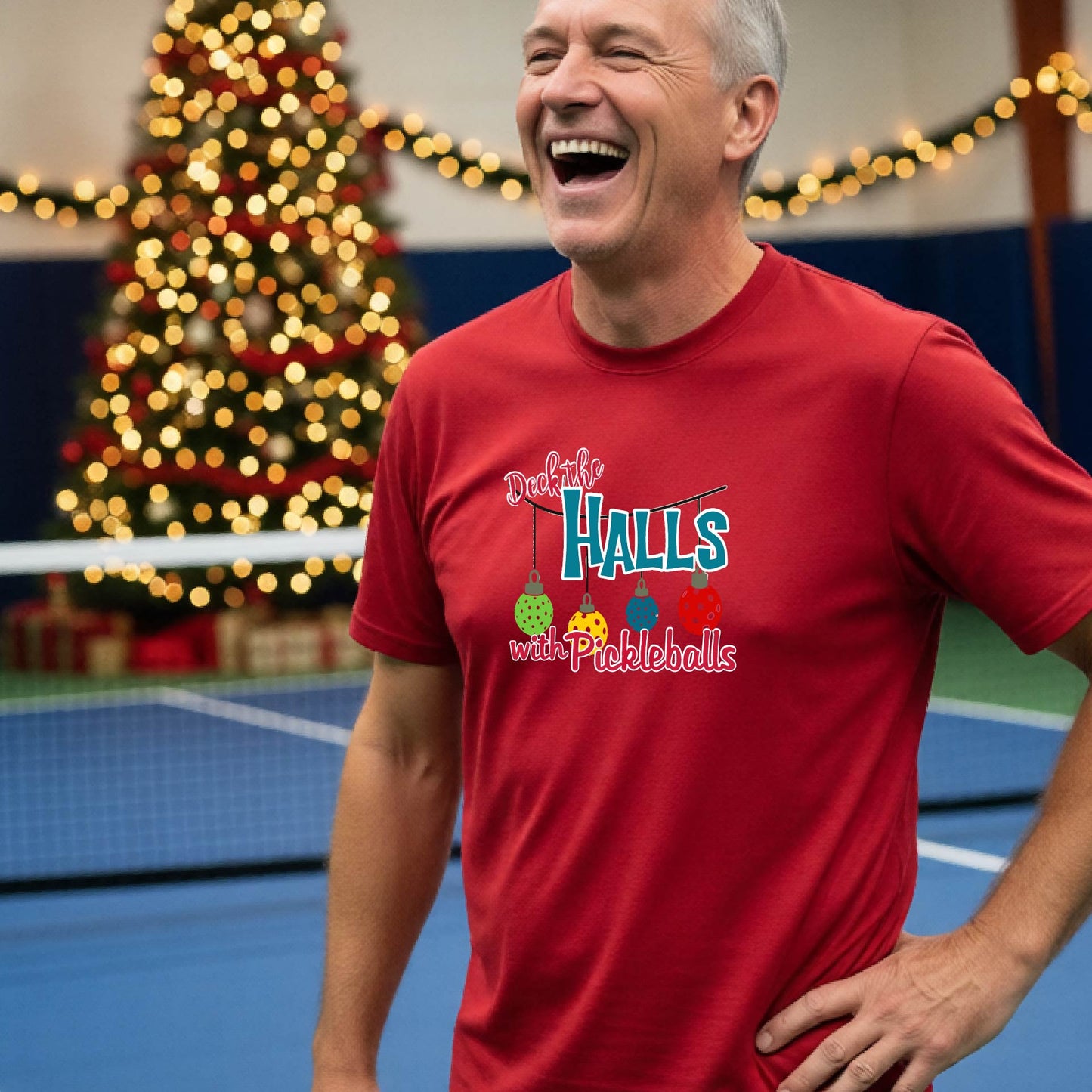 A man wearing a red graphic t-shirt with a 'Deck the  Halls with Pickleballs design, standing in front of a blurred Christmas tree and pickleball court. 