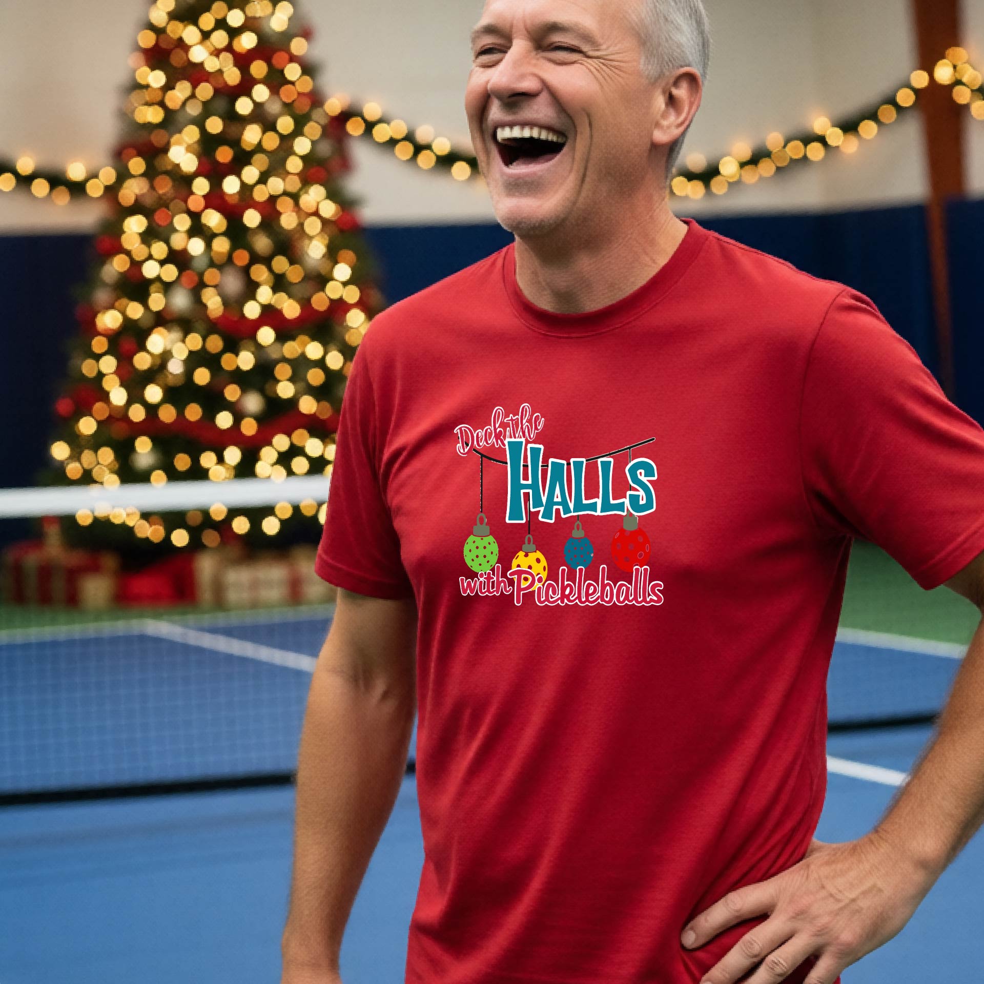 A man wearing a red graphic t-shirt with a 'Deck the  Halls with Pickleballs design, standing in front of a blurred Christmas tree and pickleball court. 