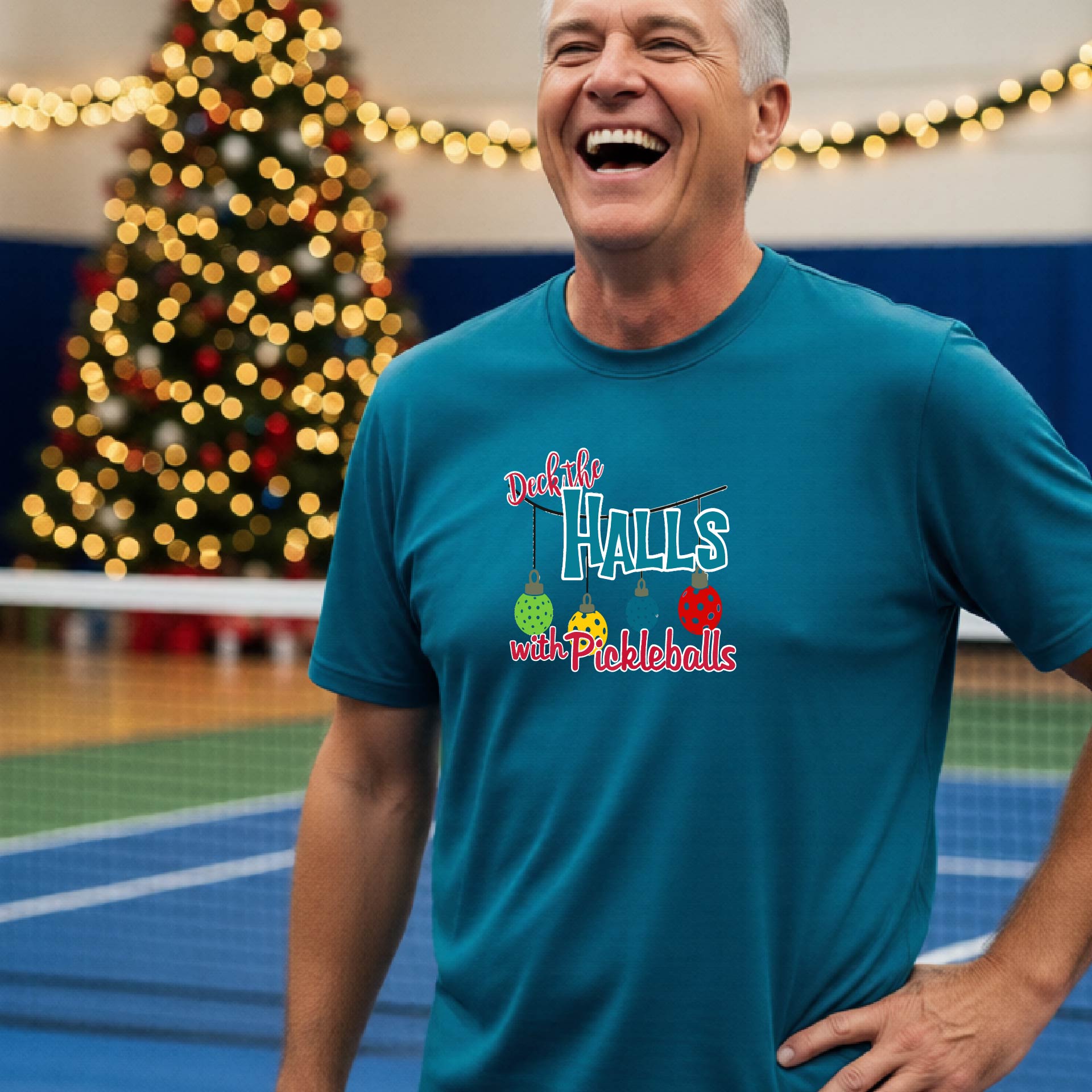 A man wearing a blue graphic t-shirt with a 'Deck the  Halls with Pickleballs design, standing in front of a blurred Christmas tree and pickleball court. 