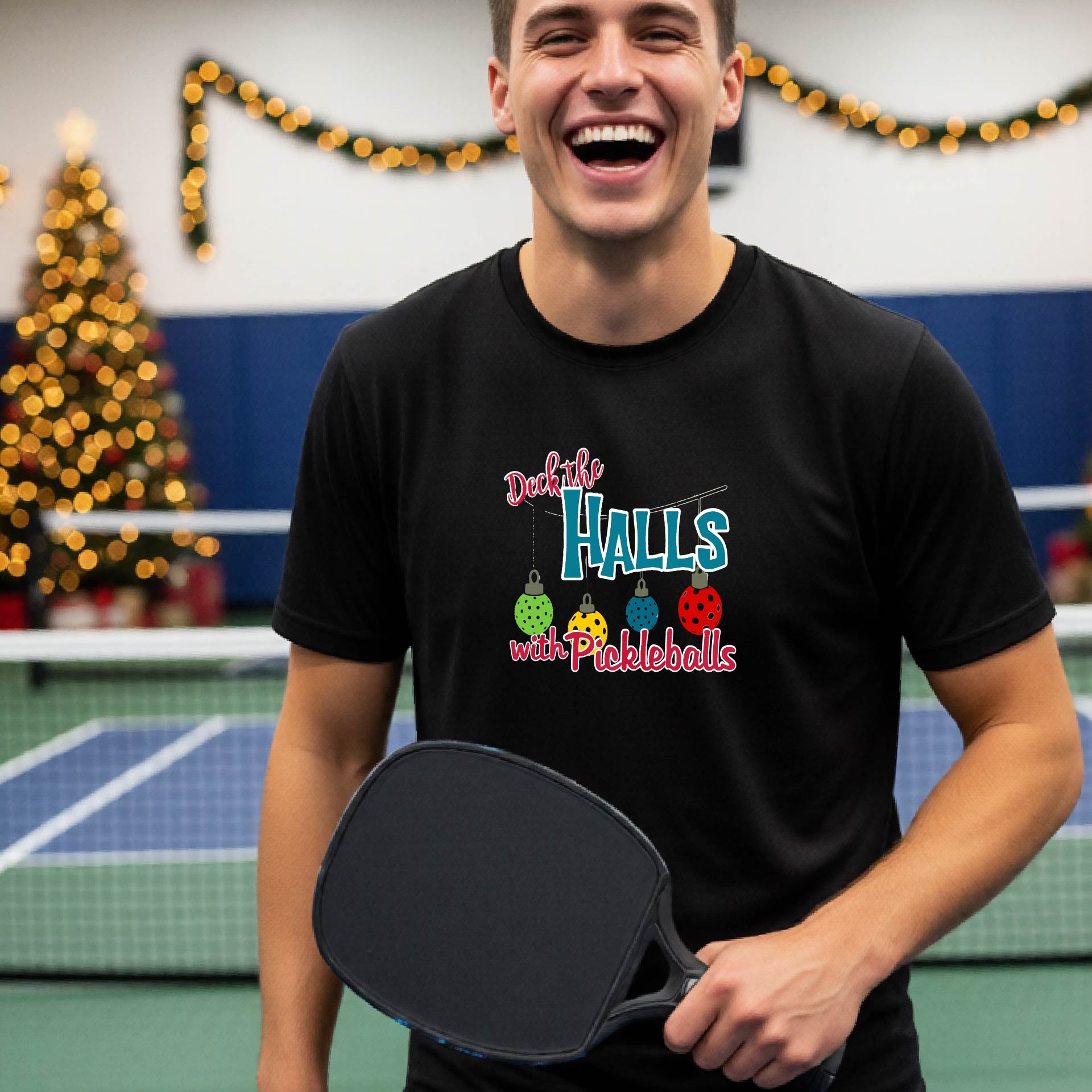A man wearing a black graphic t-shirt with a 'Deck the  Halls with Pickleballs design, standing in front of a blurred Christmas tree and pickleball court. 