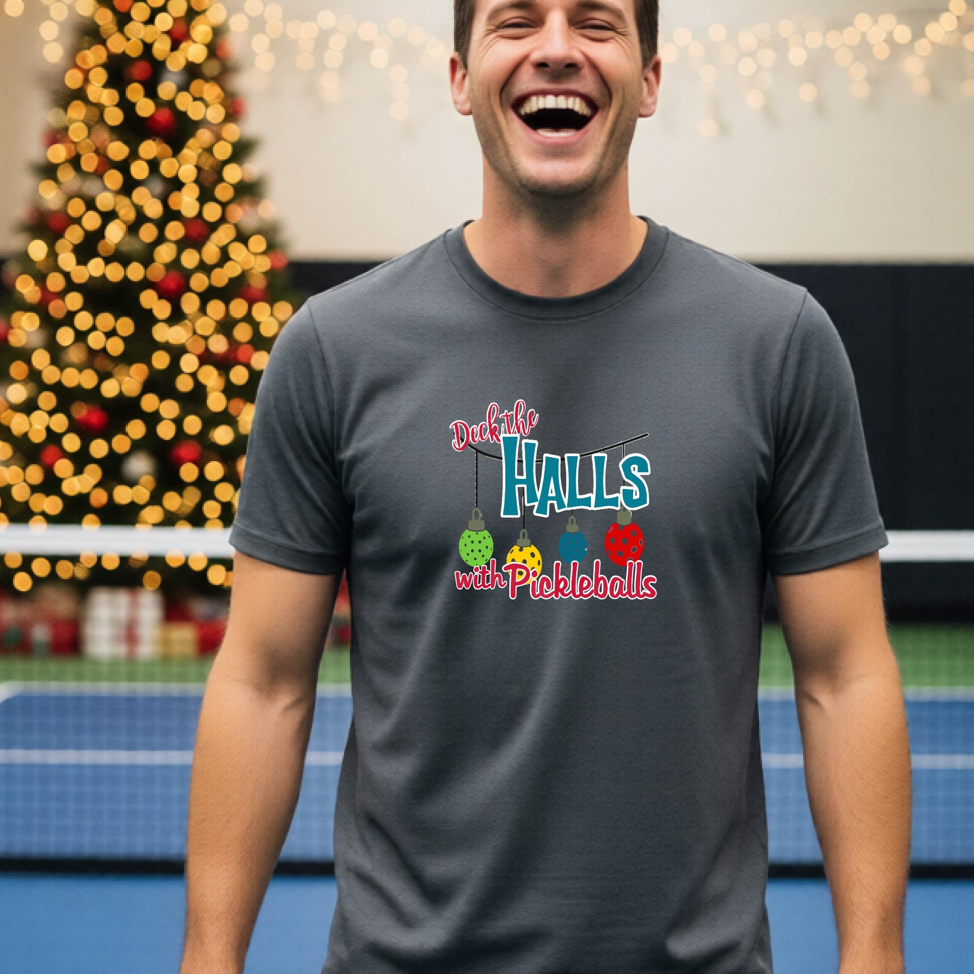A man wearing a dark gray graphic t-shirt with a 'Deck the  Halls with Pickleballs design, standing in front of a blurred Christmas tree and pickleball court. 