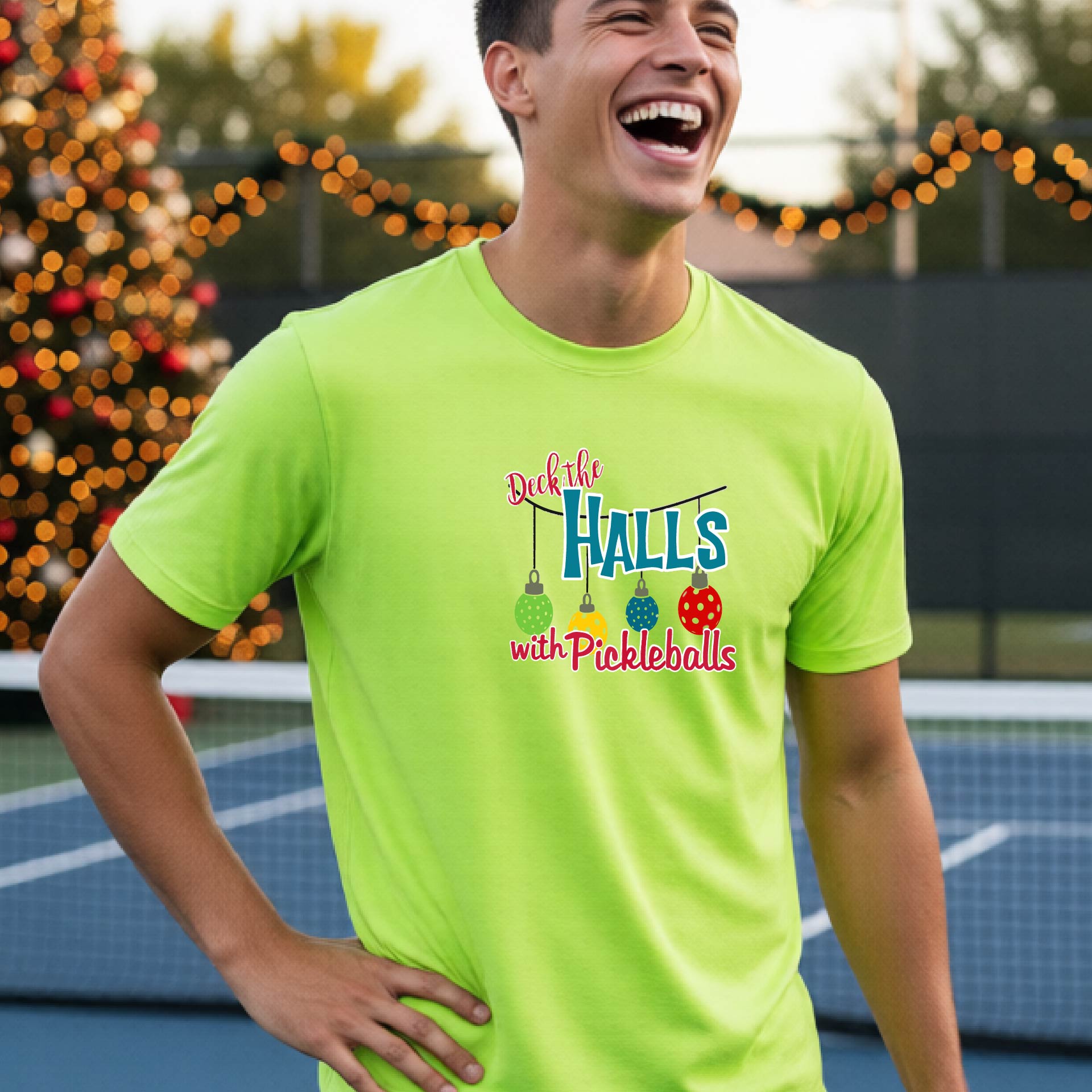 A man wearing a lime green graphic t-shirt with a 'Deck the  Halls with Pickleballs' design, standing in front of a blurred Christmas tree and pickleball court. 