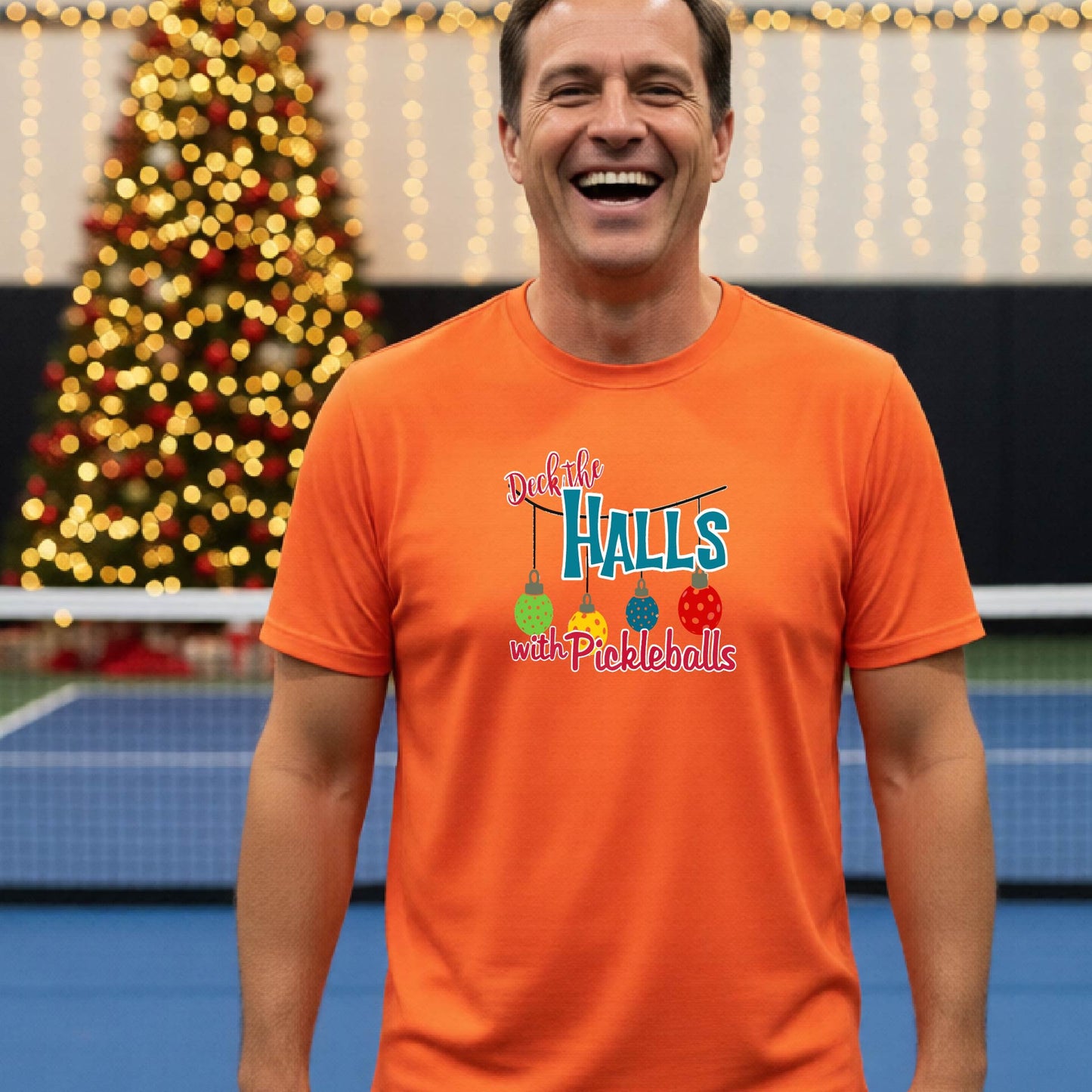 A man wearing a bright orange graphic t-shirt with a 'Deck the  Halls with Pickleballs design, standing in front of a blurred Christmas tree and pickleball court. 