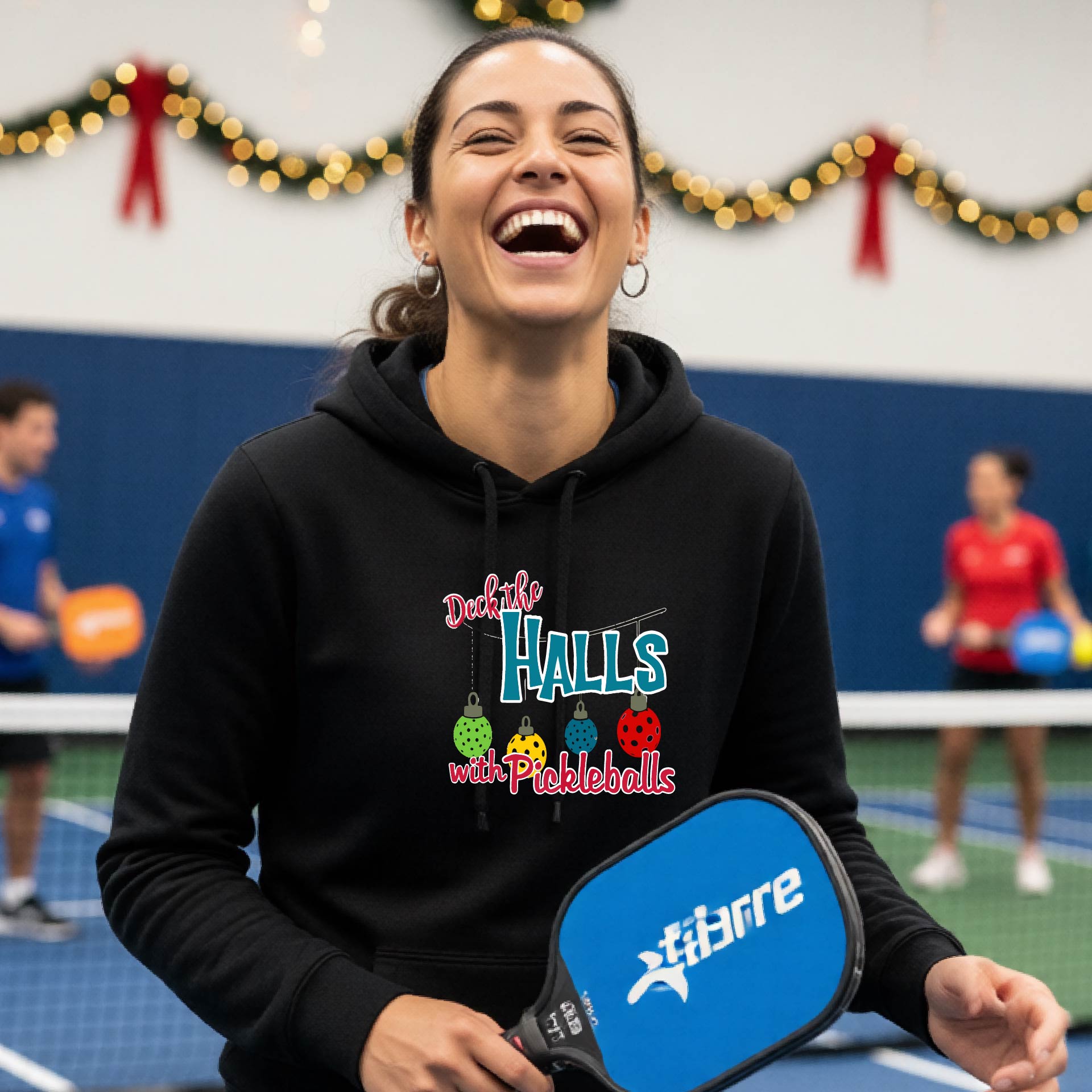 A black hooded sweatshirt featuring a graphic print that reads "Deck the Halls with Picleballs" . The backgro0und is Christmas themed. 