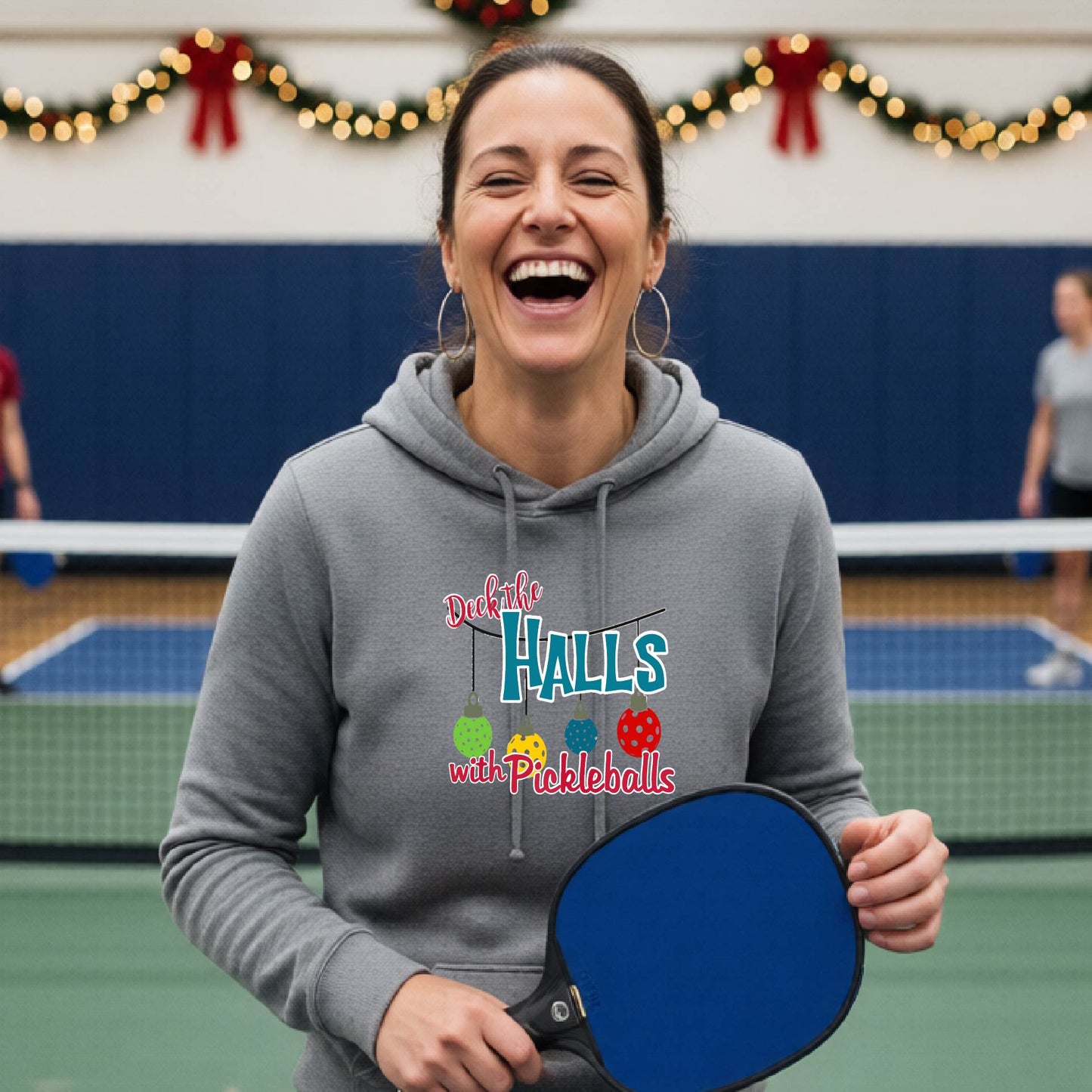 A gray hooded sweatshirt featuring a graphic print that reads "Deck the Halls with Picleballs" . The backgro0und is Christmas themed. 