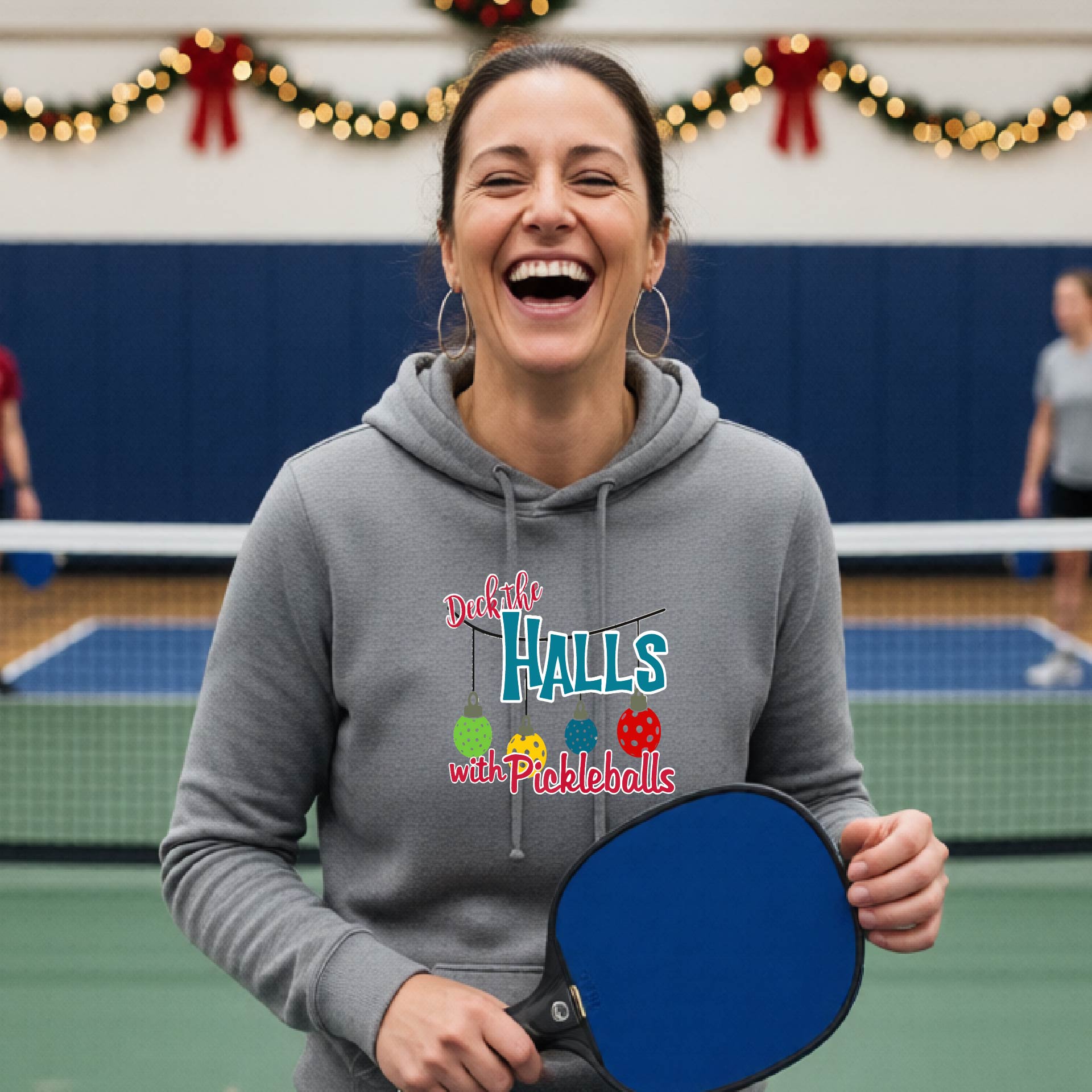 A gray hooded sweatshirt featuring a graphic print that reads "Deck the Halls with Picleballs" . The backgro0und is Christmas themed. 