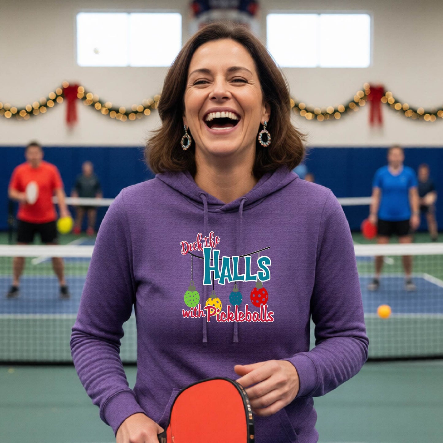 A purple hooded sweatshirt featuring a graphic print that reads "Deck the Halls with Picleballs" . The backgro0und is Christmas themed. 