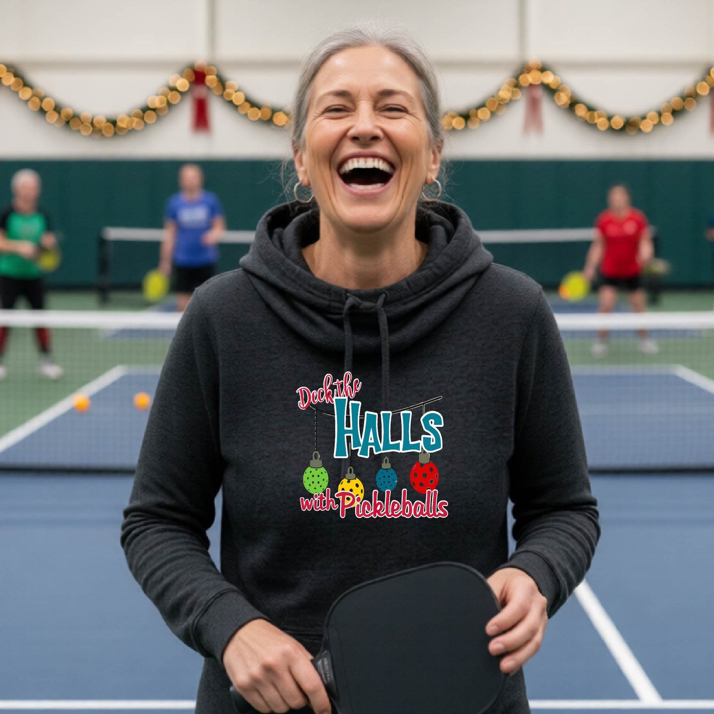 A black cowl-neck hooded sweatshirt featuring a graphic print that reads 'Deck the Halls with Pickleballs" in red, white, and teal text. The design includes four colorful pickleball ornaments hanging from the text on thin black lines.