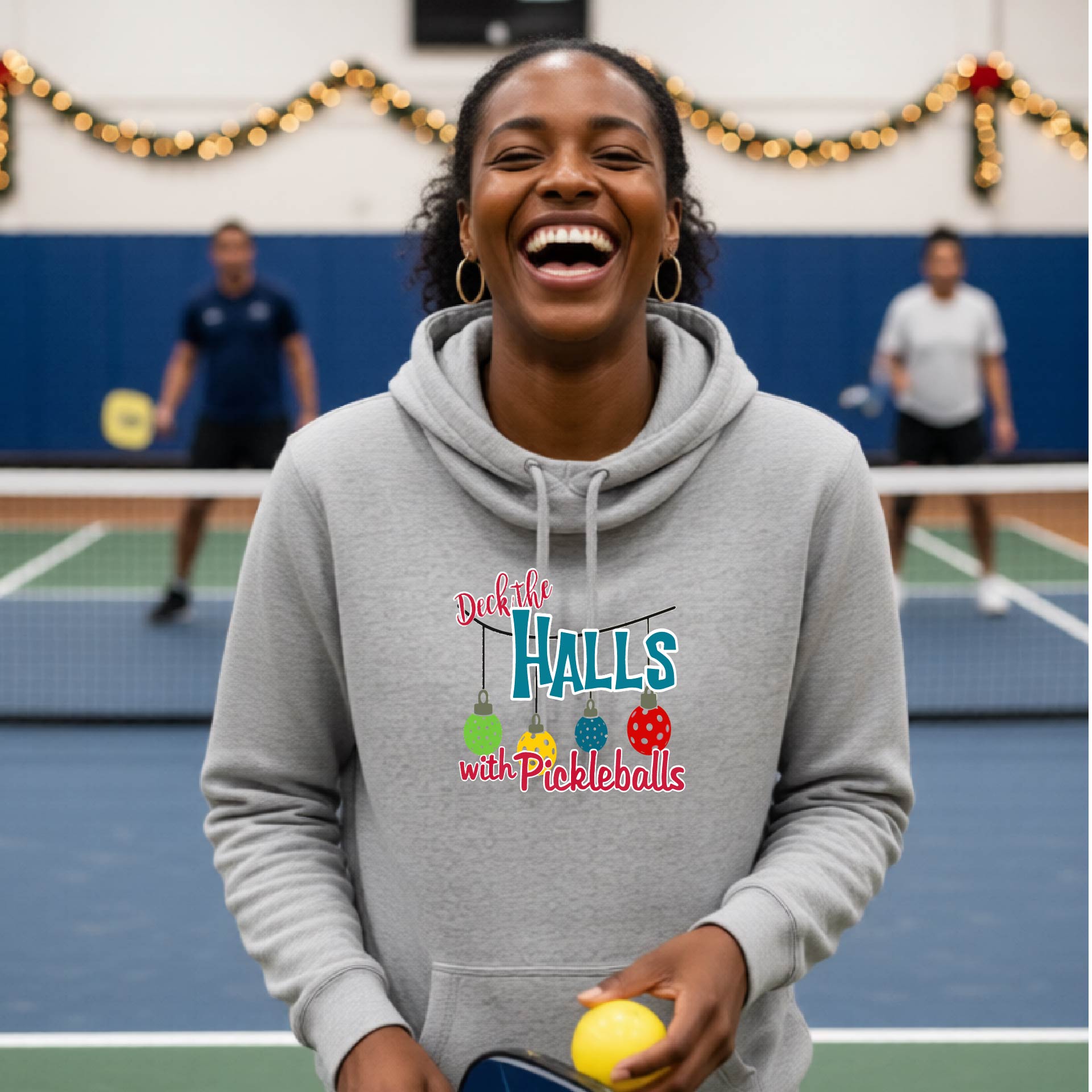 A gray cowl-neck hooded sweatshirt featuring a graphic print that reads 'Deck the Halls with Pickleballs" in red, white, and teal text. The design includes four colorful pickleball ornaments hanging from the text on thin black lines.