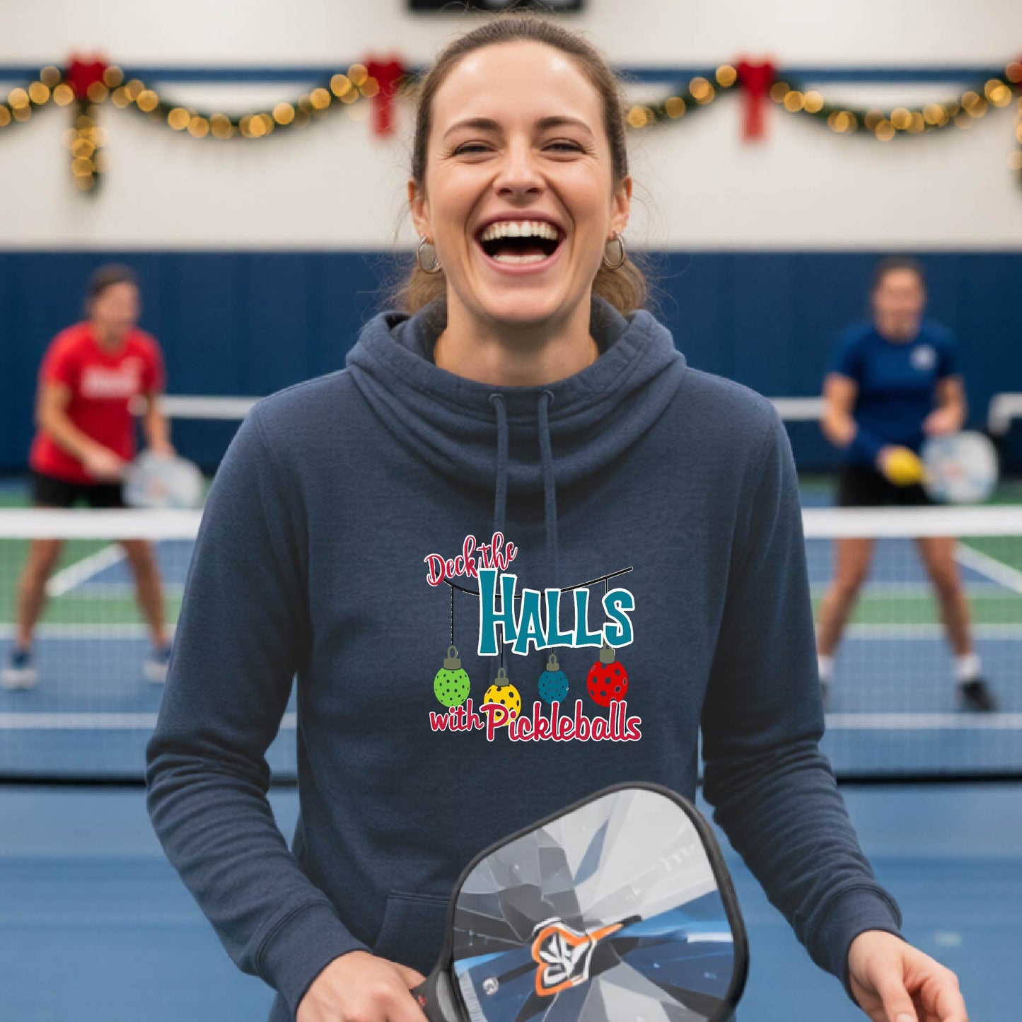 A navy cowl-neck hooded sweatshirt featuring a graphic print that reads 'Deck the Halls with Pickleballs" in red, white, and teal text. The design includes four colorful pickleball ornaments hanging from the text on thin black lines.