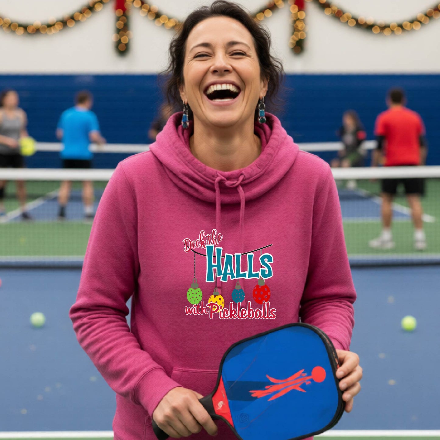A pink cowl-neck hooded sweatshirt featuring a graphic print that reads 'Deck the Halls with Pickleballs" in red, white, and teal text. The design includes four colorful pickleball ornaments hanging from the text on thin black lines.