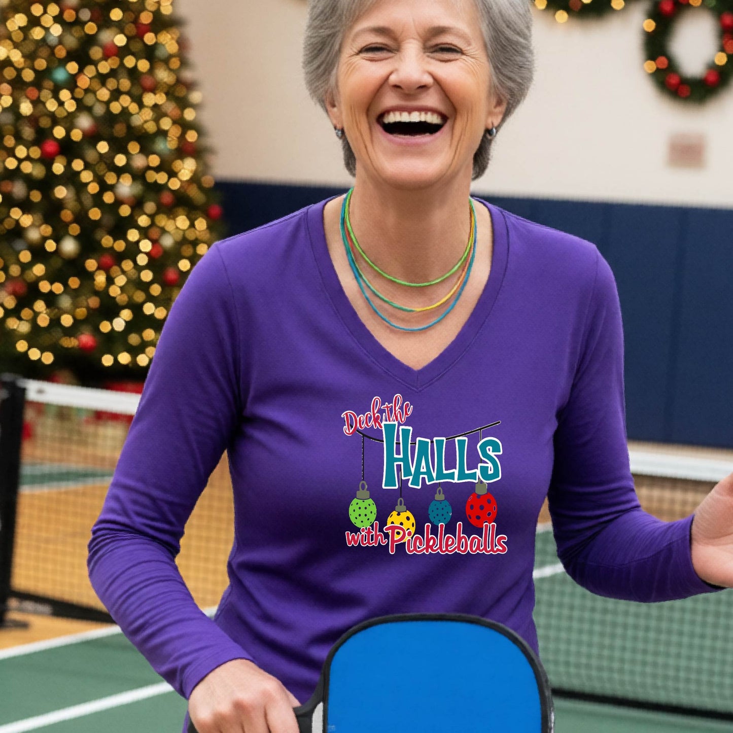A women's purple  long-sleeve v-neck shirt featuring a pickleball graphic with text "DECK THE HALLS WITH PICKLEBALLS"  and  Christmas themed background. 