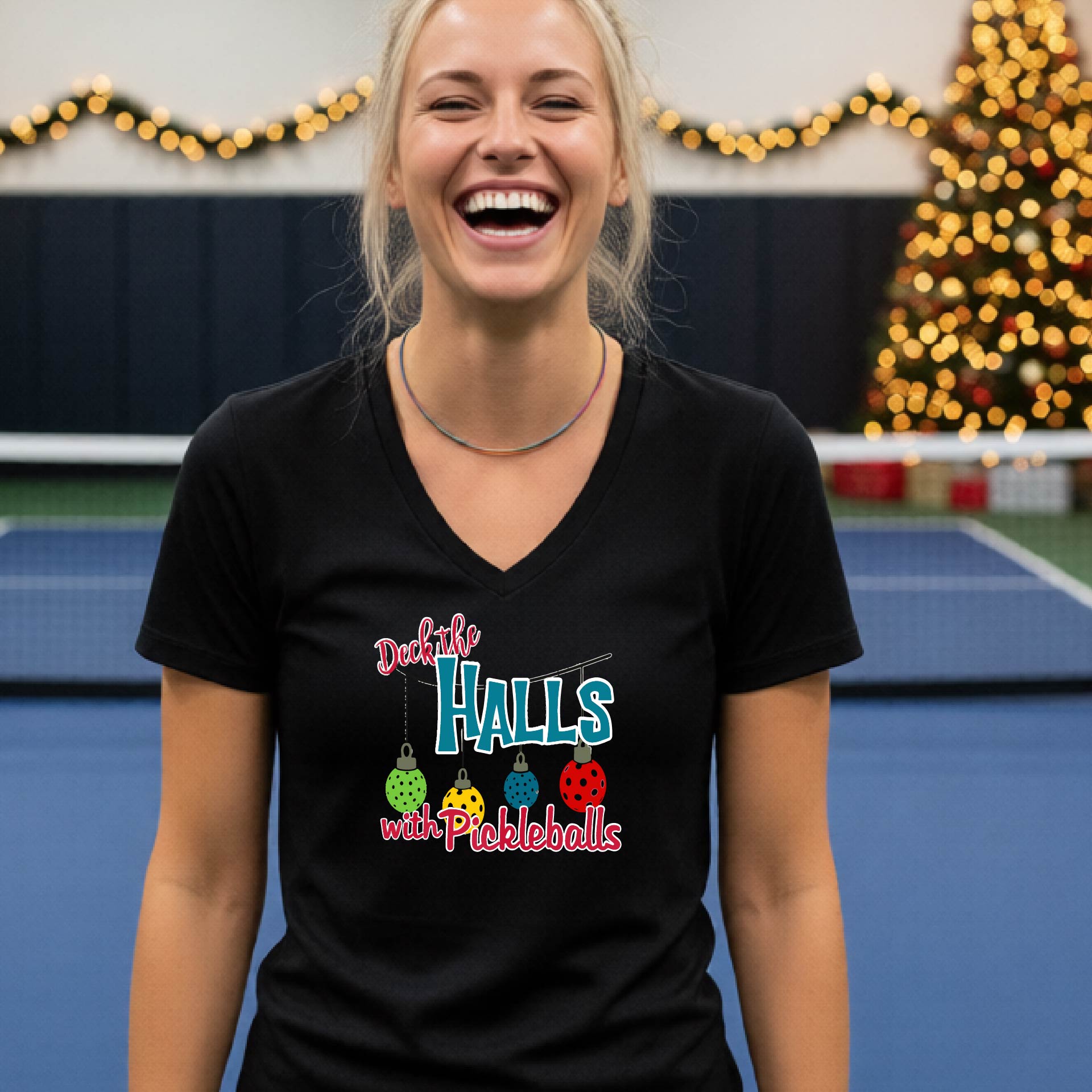 A woman laughing on an indoor pickleball court wearing a black V-neck t-shirt with a colorful graphic that reads 'Deck the Halls with Pickleballs' and shows three hanging pickleballs. A Christmas tree and holiday decorations are blurred in the background.