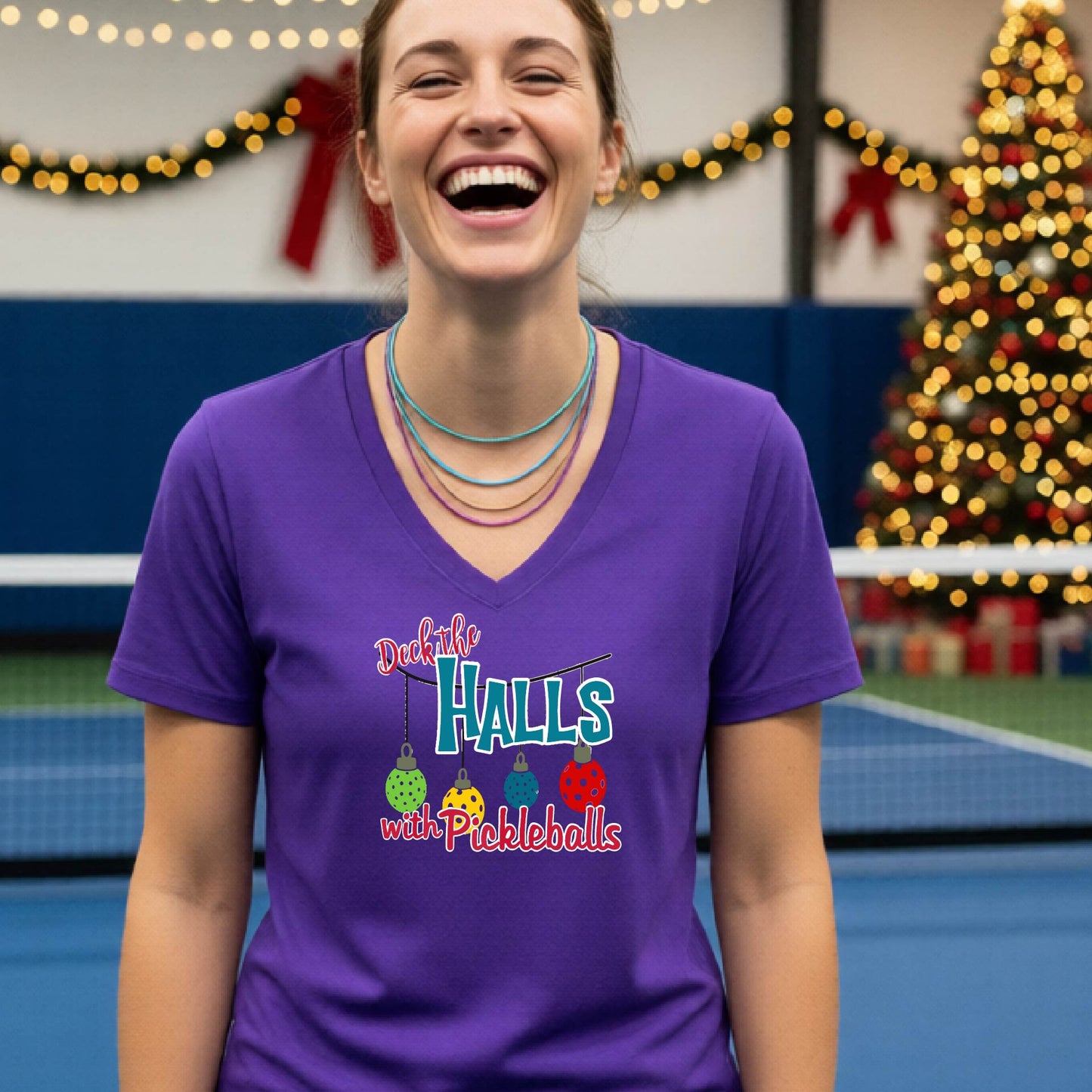 A woman laughing on an indoor pickleball court wearing a purple V-neck t-shirt with a colorful graphic that reads 'Deck the Halls with Pickleballs' and shows three hanging pickleballs. A Christmas tree and holiday decorations are blurred in the background.