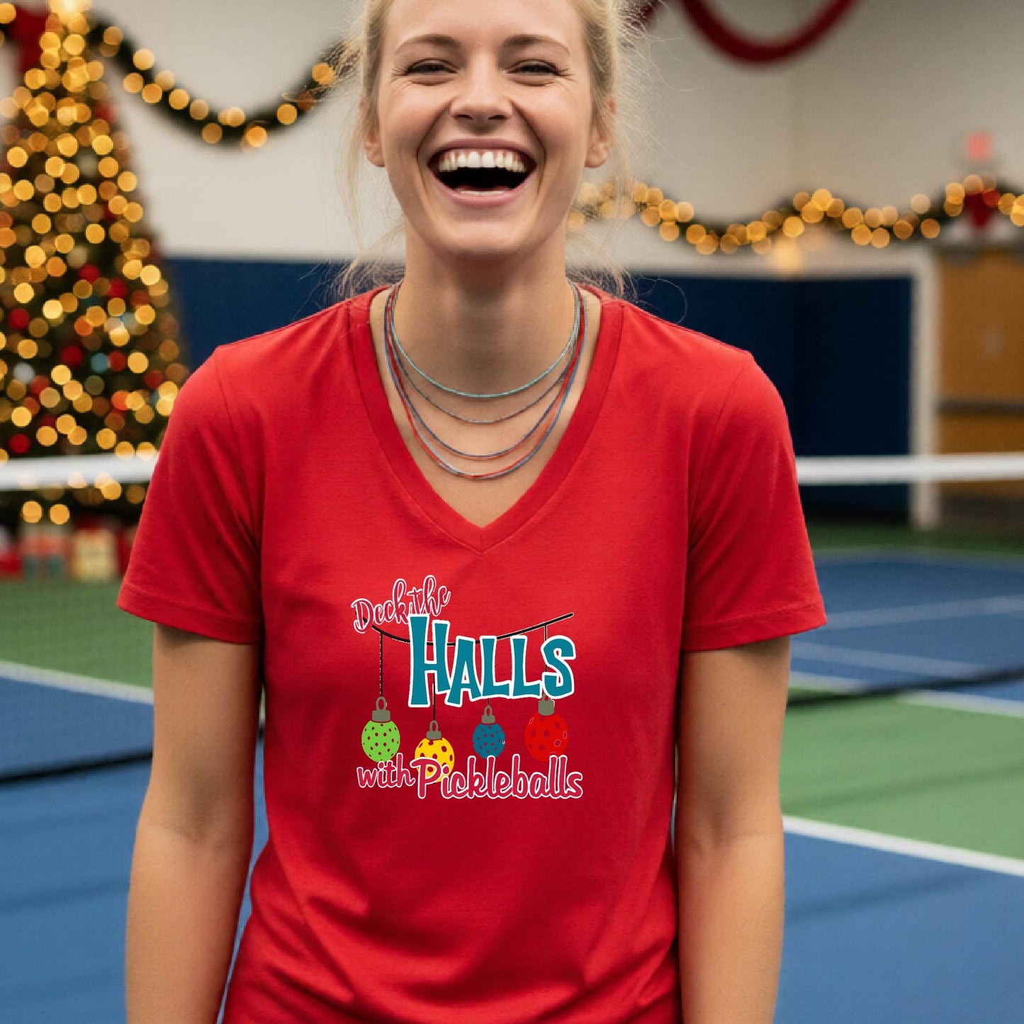 A woman laughing on an indoor pickleball court wearing a red V-neck t-shirt with a colorful graphic that reads 'Deck the Halls with Pickleballs' and shows three hanging pickleballs. A Christmas tree and holiday decorations are blurred in the background.