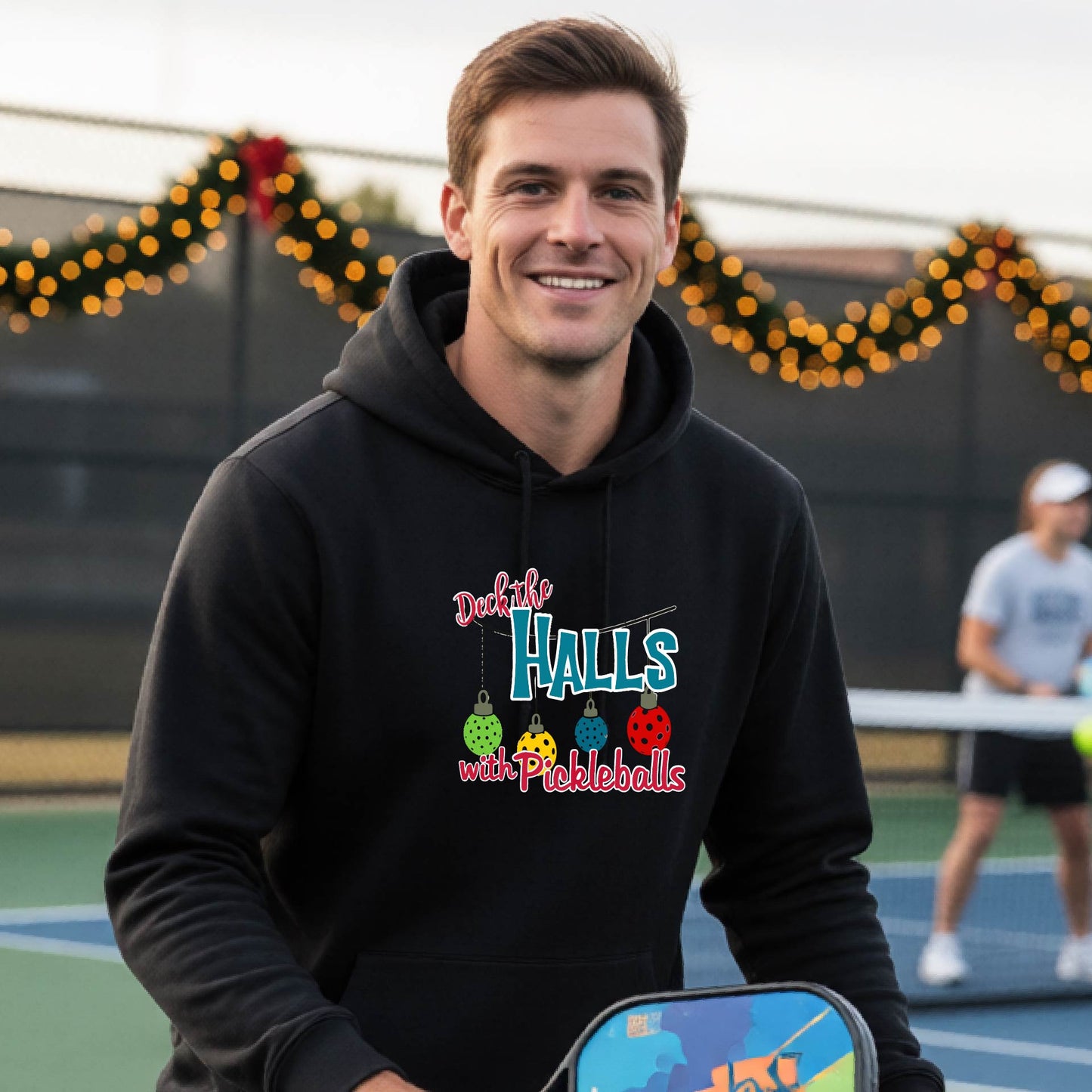 A smiling man wearing a black hoodie stands on a pickleball court. The hoodie has a festive graphic that reads 'Deck the Halls with Pickleballs' with Christmas pickleball ornaments.