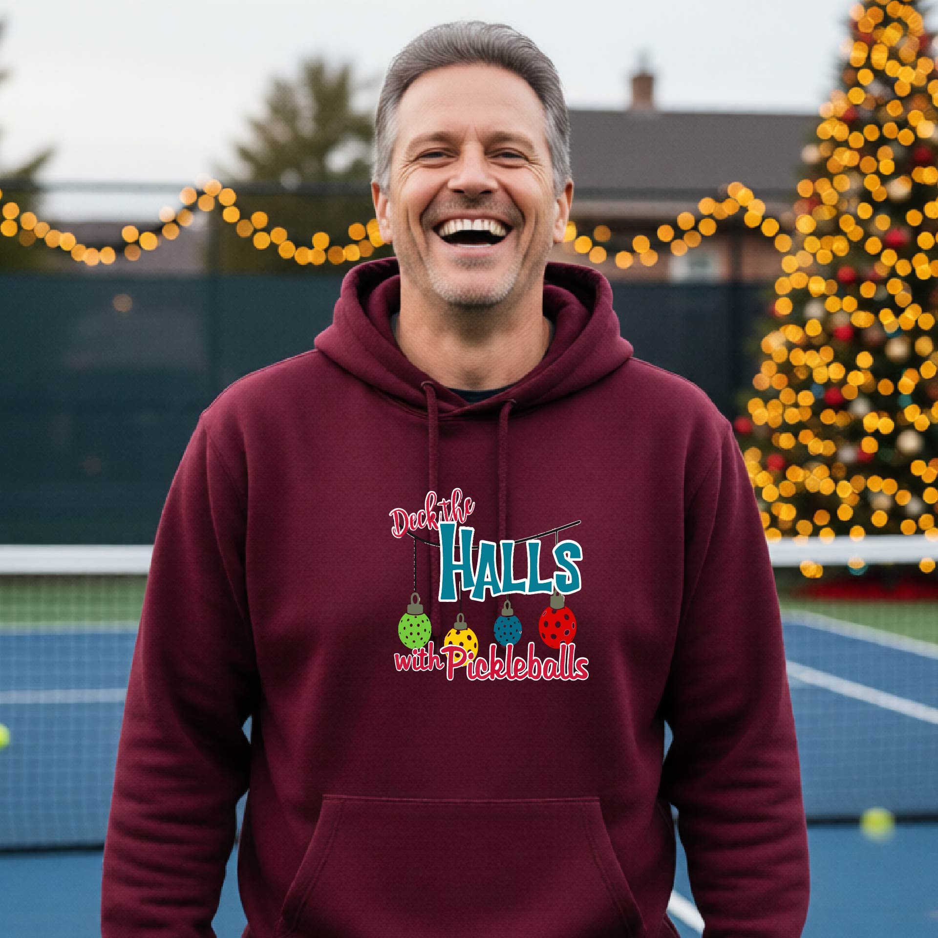 A smiling man wearing a aroonhoodie stands on a pickleball court. The hoodie has a festive graphic that reads 'Deck the Halls with Pickleballs' with Christmas pickleball ornaments.
