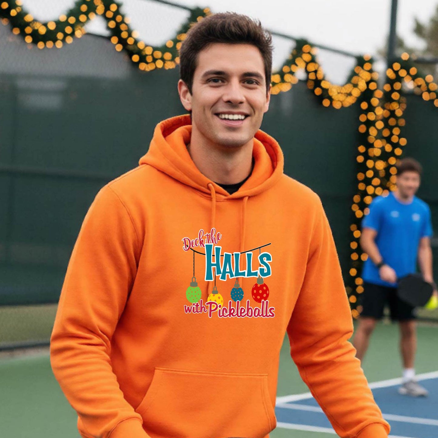 A smiling man wearing a bright orange hoodie stands on a pickleball court. The hoodie has a festive graphic that reads 'Deck the Halls with Pickleballs' with Christmas pickleball ornaments.