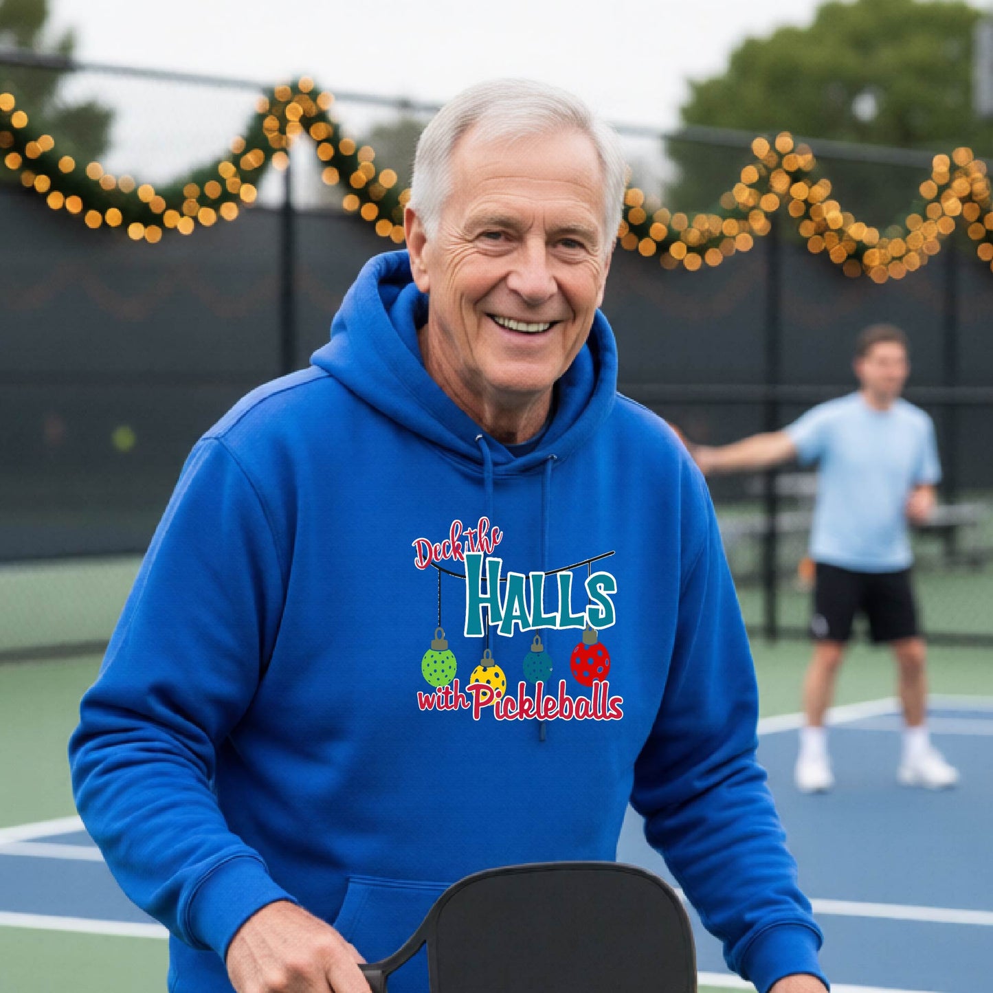 A smiling man wearing a bright royal blue hoodie stands on a pickleball court. The hoodie has a festive graphic that reads 'Deck the Halls with Pickleballs' with Christmas pickleball ornaments.