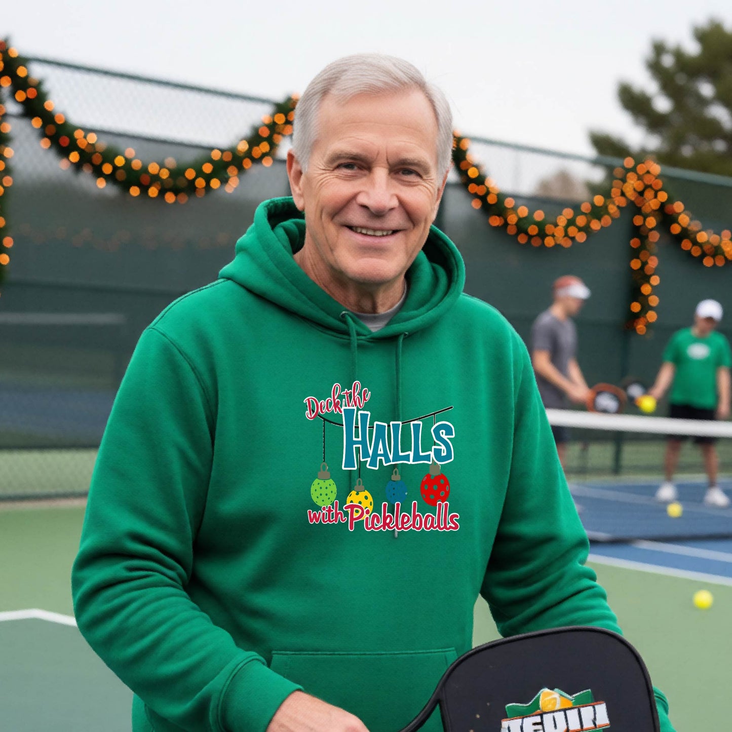 A smiling man wearing a bright green hoodie stands on a pickleball court. The hoodie has a festive graphic that reads 'Deck the Halls with Pickleballs' with Christmas pickleball ornaments.