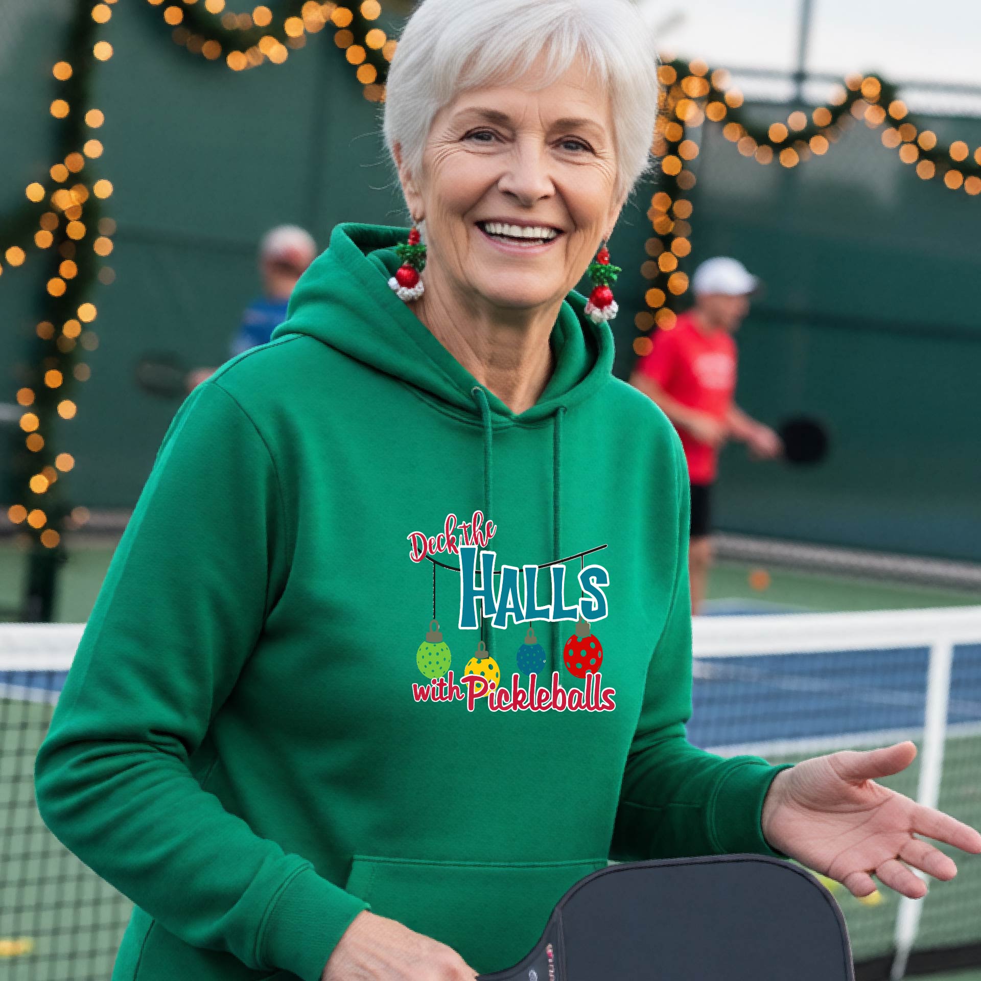 A smiling woman stands on a pickleball court. The hoodie has a festive graphic that reads 'Deck the Halls with Pickleballs' with Christmas pickleball ornaments.