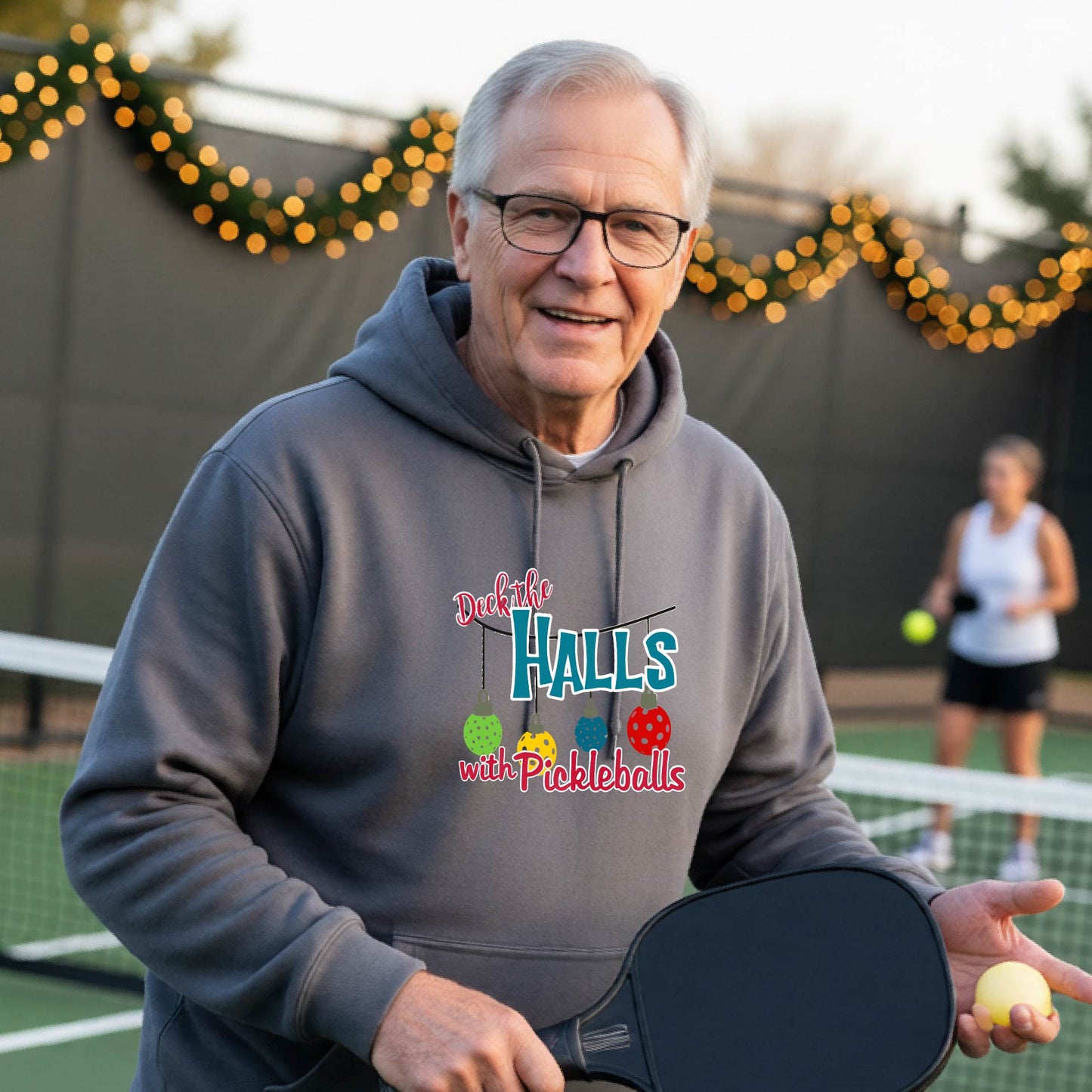 A smiling man wearing a grary hoodie stands on a pickleball court. The hoodie has a festive graphic that reads 'Deck the Halls with Pickleballs' with Christmas pickleball ornaments.
