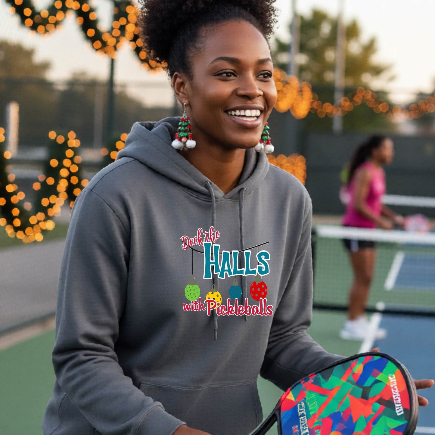 A smiling woman wearing a grey hoodie stands on a pickleball court. The hoodie has a festive graphic that reads 'Deck the Halls with Pickleballs' with Christmas pickleball ornaments.