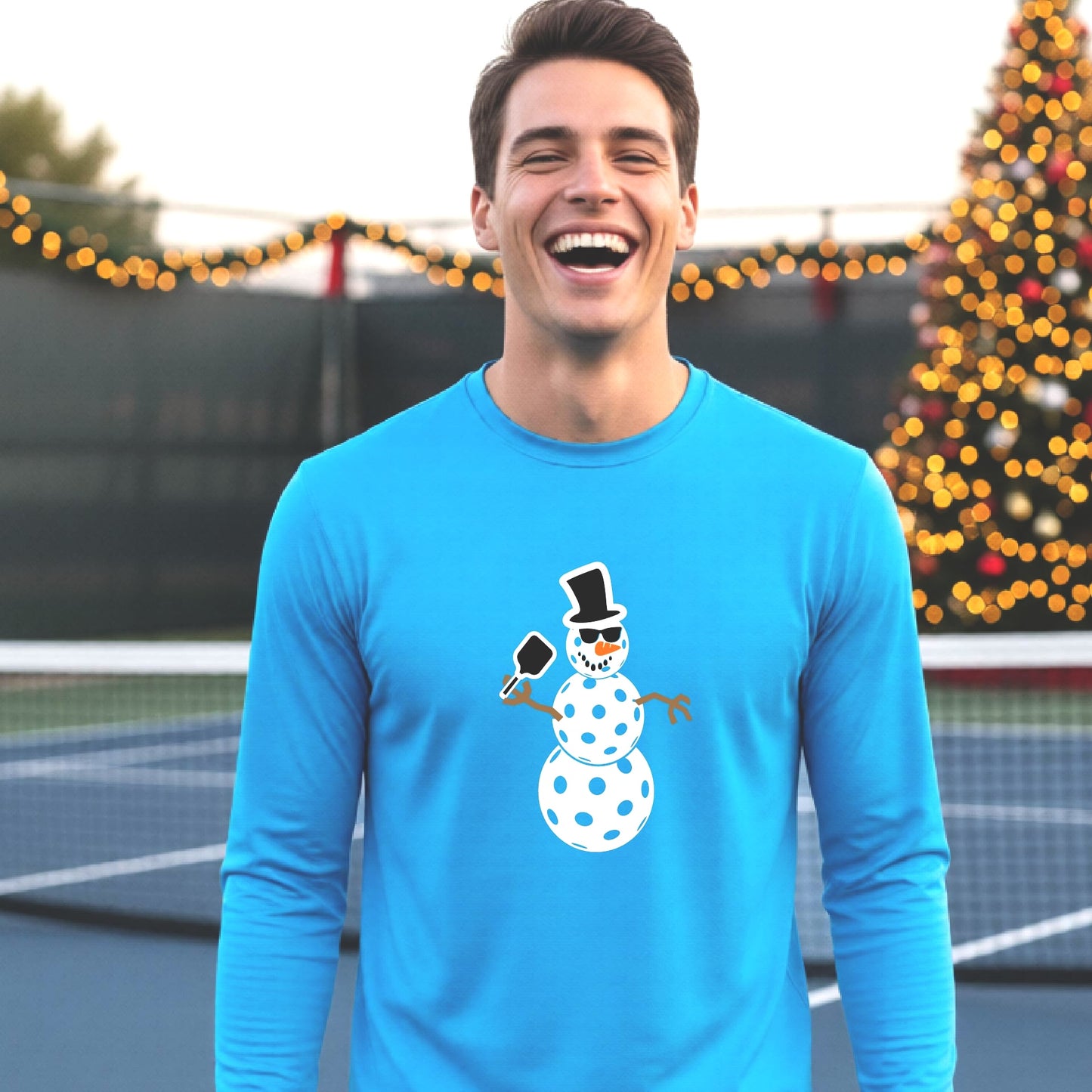 A smiling man wearing a bright blue long-sleeve shirt with a white pickleball snowman graphic on the chest, standing in front of a blurred Christmas tree with warm lights.
