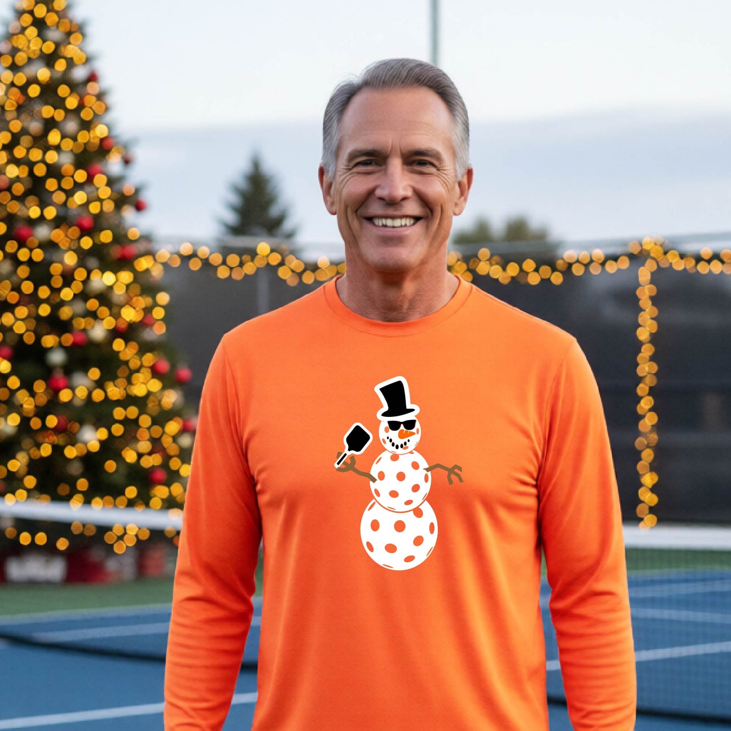 A smiling man wearing a bright orange long-sleeve shirt with a white pickleball snowman graphic on the chest, standing in front of a blurred Christmas tree with warm lights.