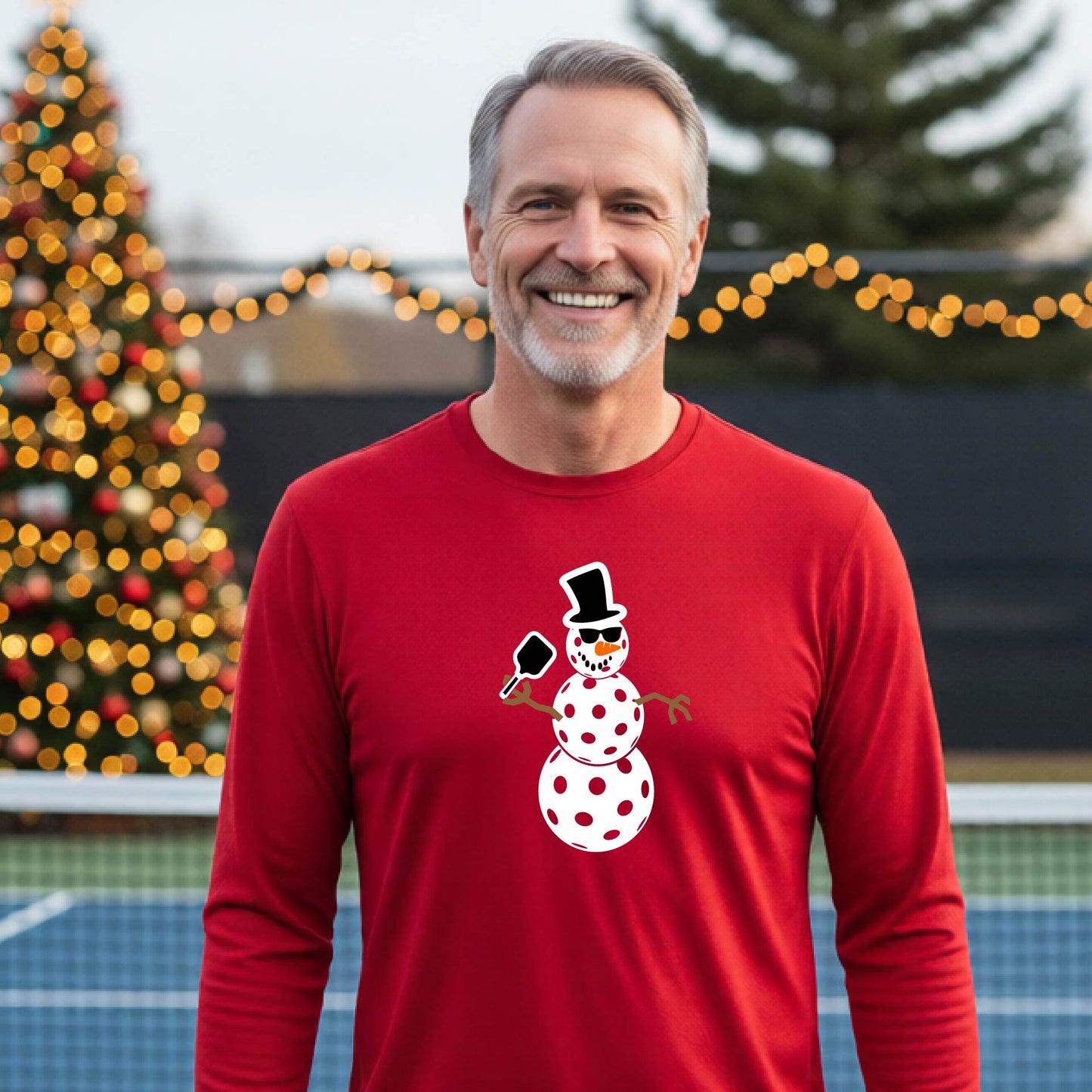 A smiling man wearing a bright red long-sleeve shirt with a white pickleball snowman graphic on the chest, standing in front of a blurred Christmas tree with warm lights.