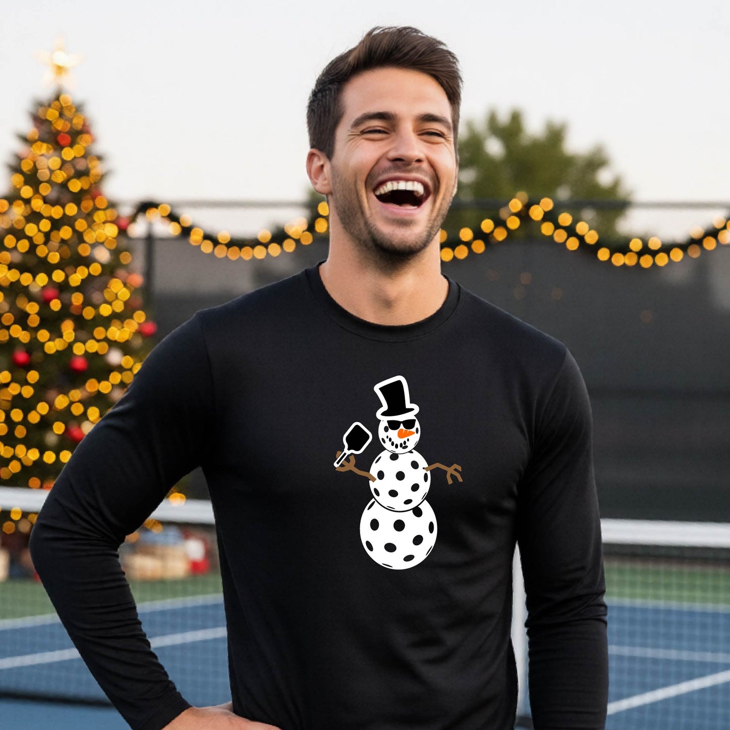 A smiling man wearing a black long-sleeve shirt with a white pickleball snowman graphic on the chest, standing in front of a blurred Christmas tree with warm lights.