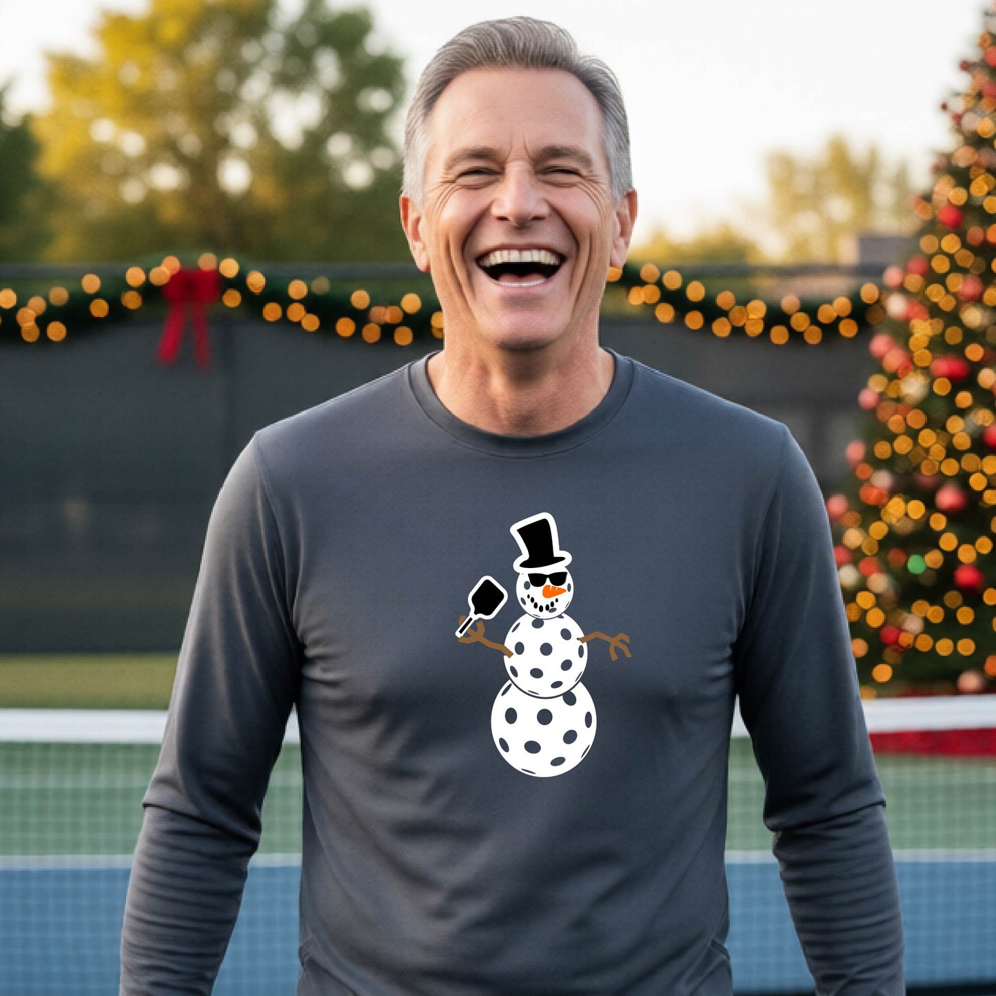 A smiling man wearing a gray long-sleeve shirt with a white pickleball snowman graphic on the chest, standing in front of a blurred Christmas tree with warm lights.