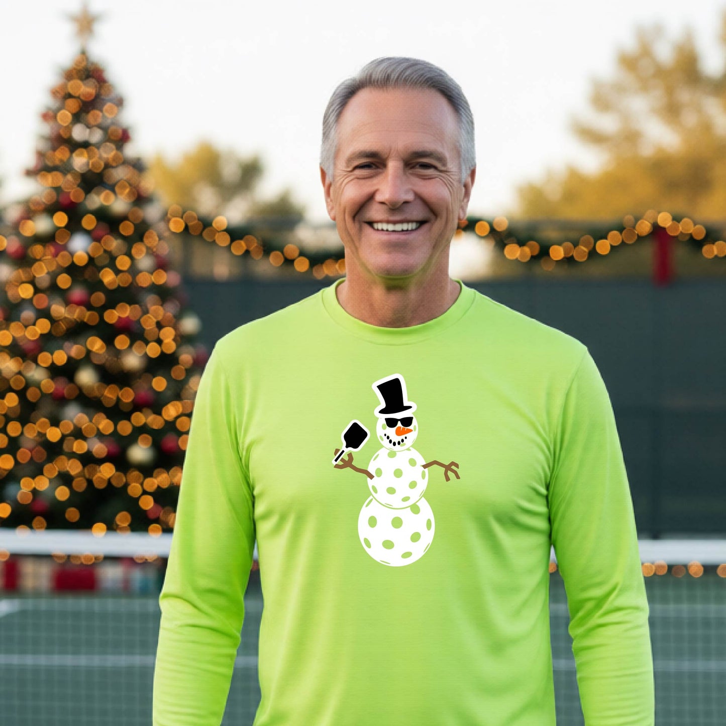 A smiling man wearing a bright lime green long-sleeve shirt with a white pickleball snowman graphic on the chest, standing in front of a blurred Christmas tree with warm lights.